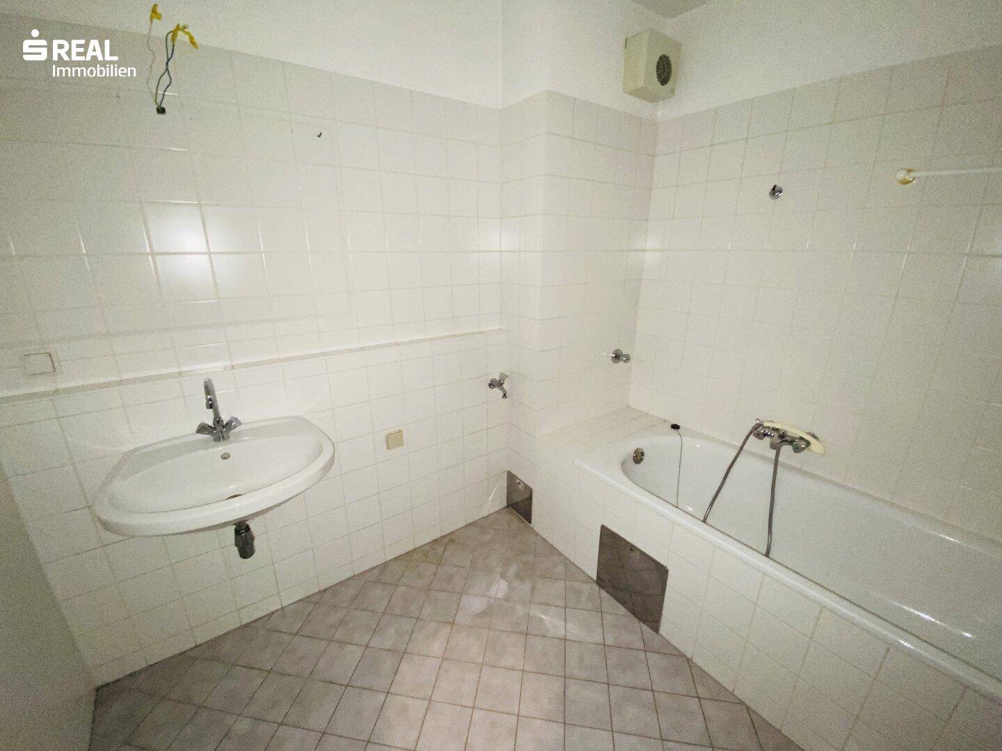Badezimmer mit weißen Fliesen, Waschbecken und Badewanne, in originalem Zustand.