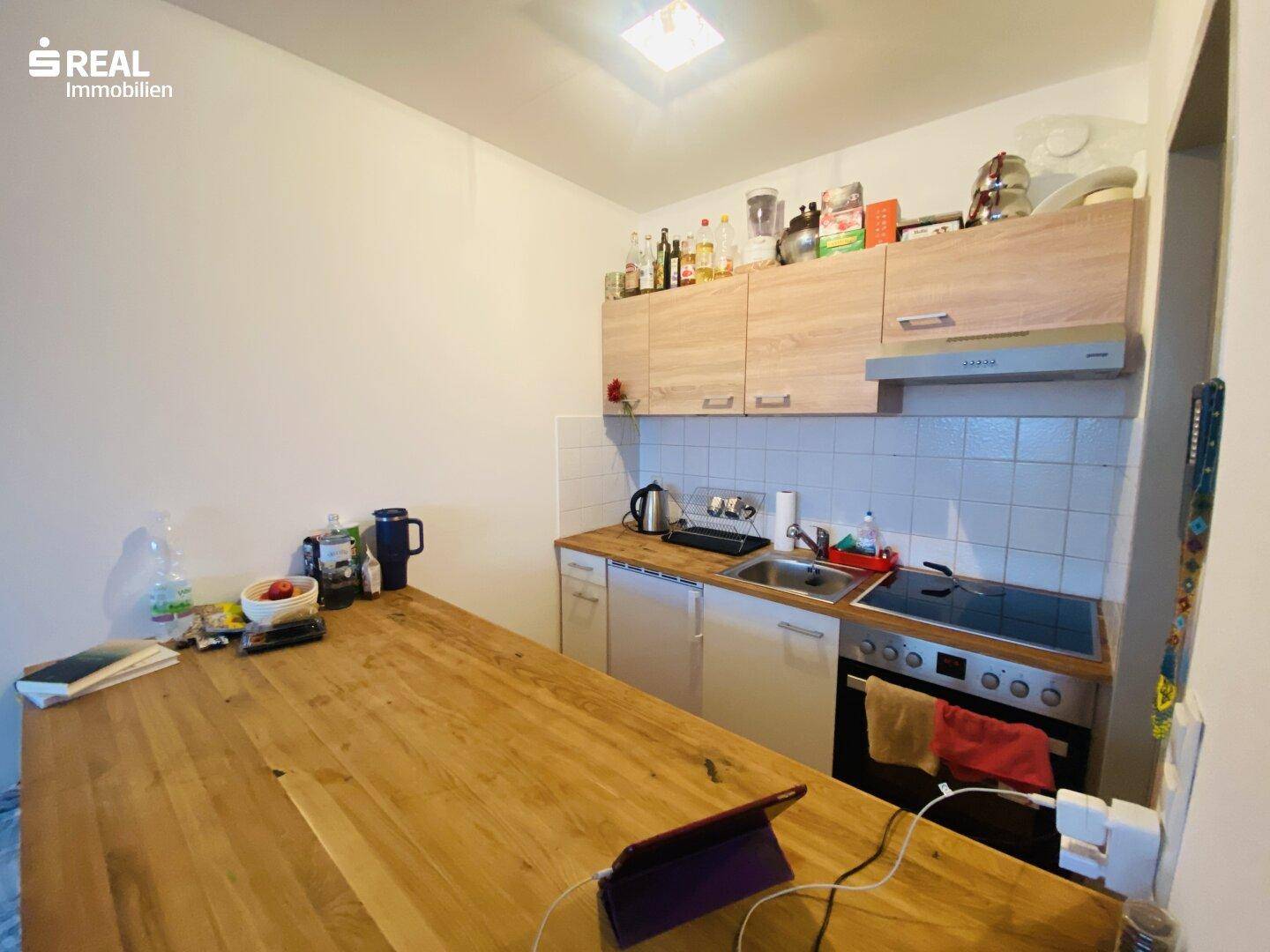 Kleine Küchenzeile mit Holz-Arbeitsplatte, Kochfeld, Backofen und weißen Fliesen als Spritzschutz.