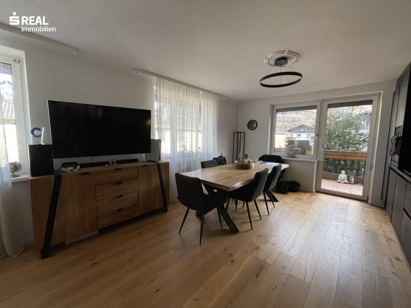 Heller Wohn- und Essbereich mit Holzboden, großem Fernseher und Zugang zum Balkon.