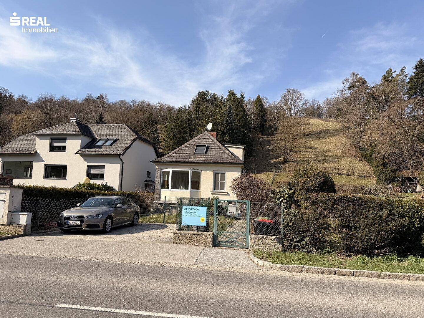 Einfamilienhaus mit Vorgarten und Parkplatz an der Straße, umgeben von grüner Natur.