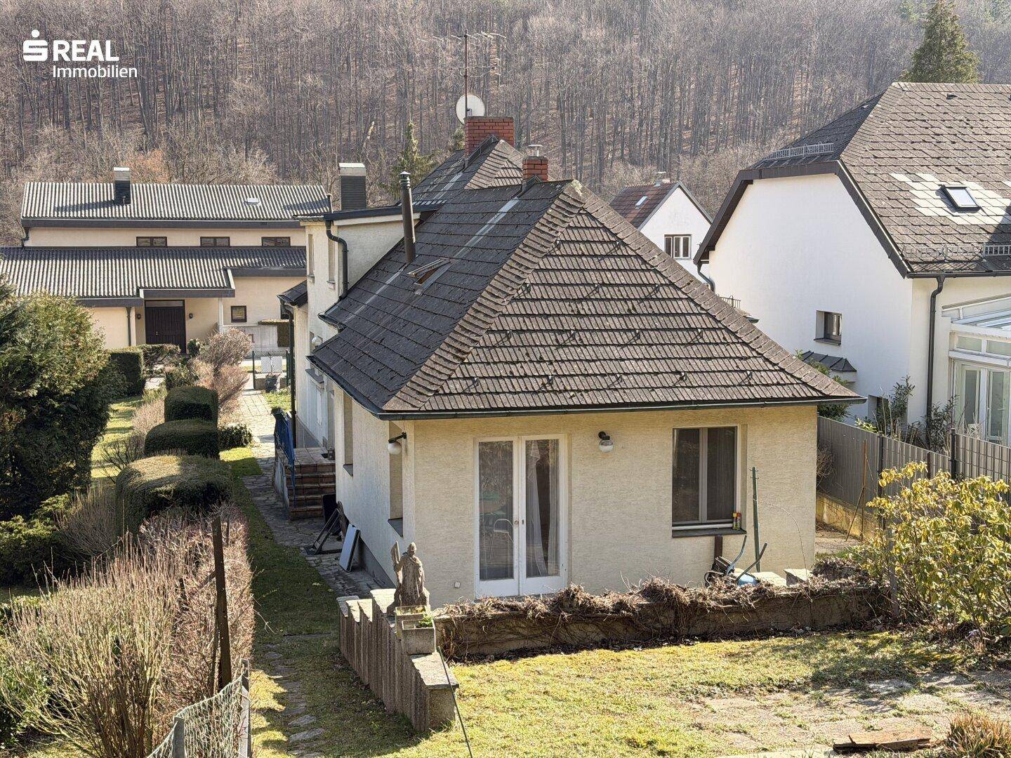 Rückansicht des Hauses mit Garten, Terrassentür und Blick auf die bewaldeten Hügel.