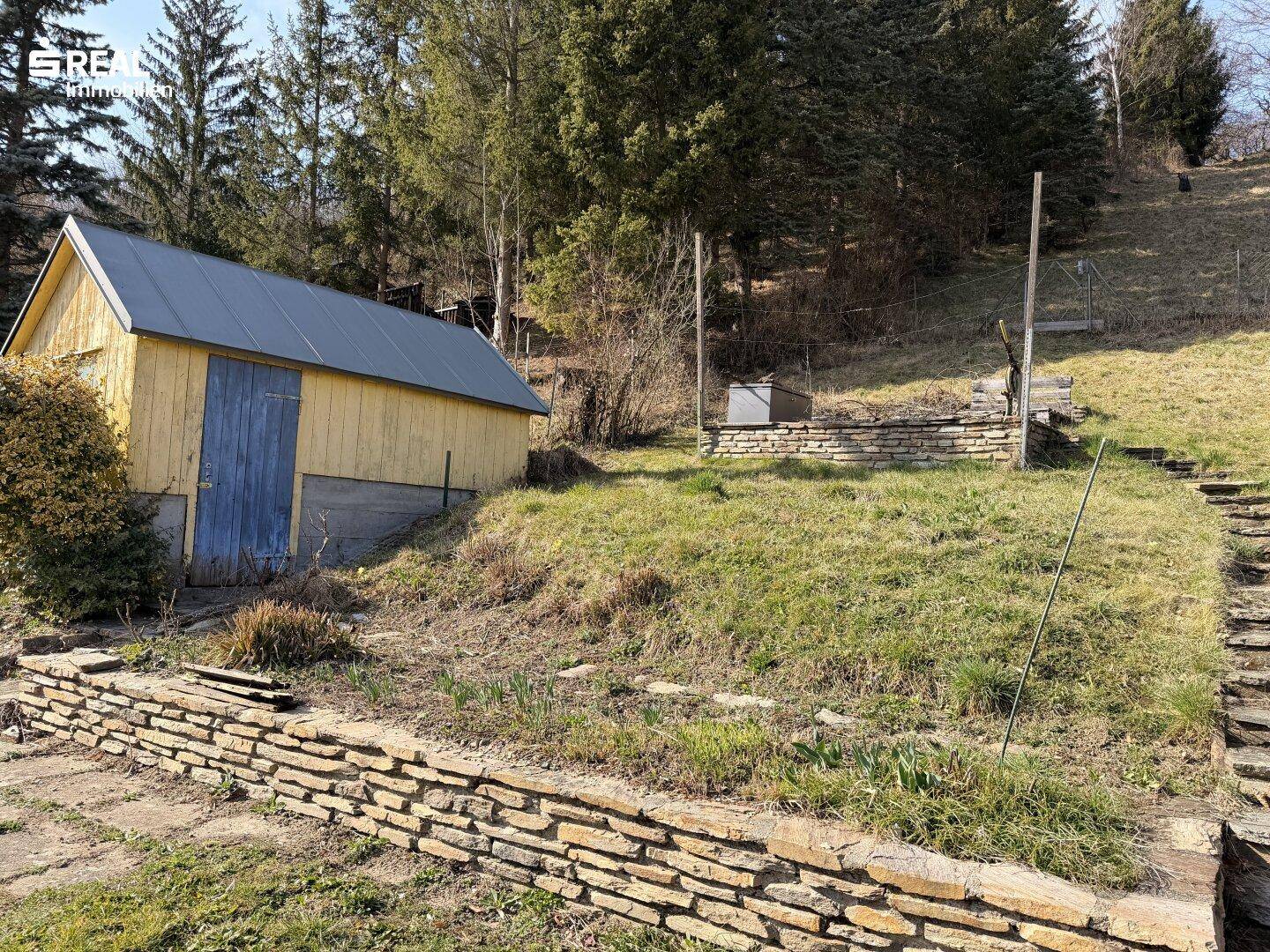 Holzschuppen und terrassierter Gartenbereich mit Steinmauern und viel Grün am Hang.