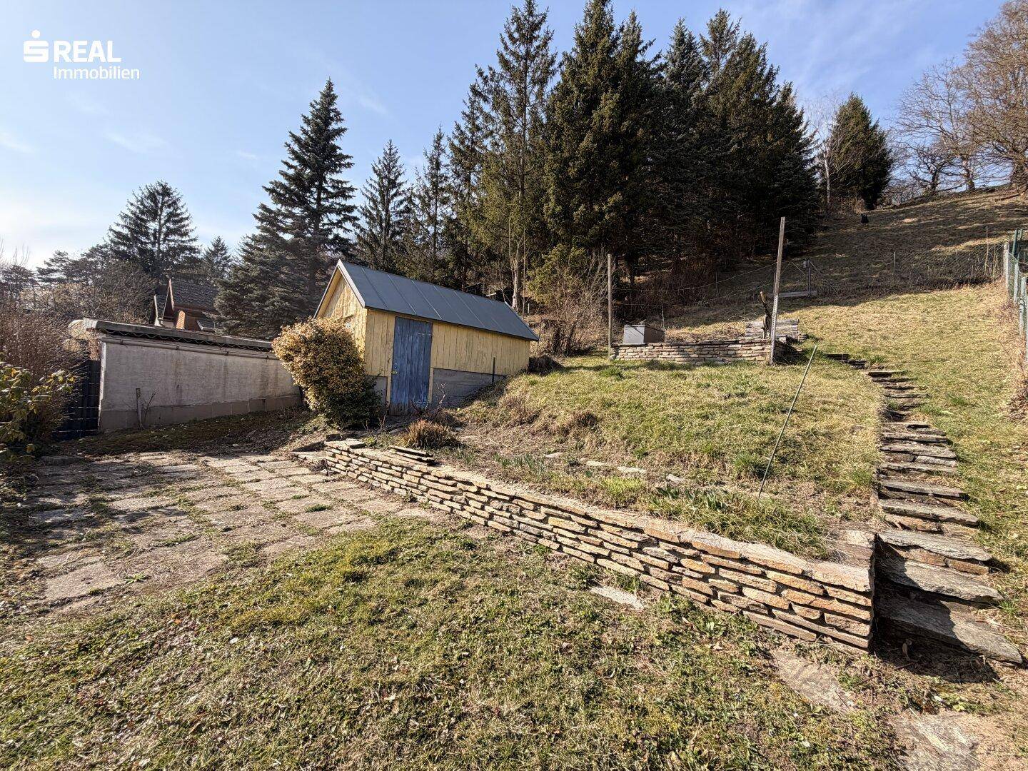 Terrassierter Gartenbereich mit Steinmauern, Holzschuppen und Zugang zu weiteren Flächen.