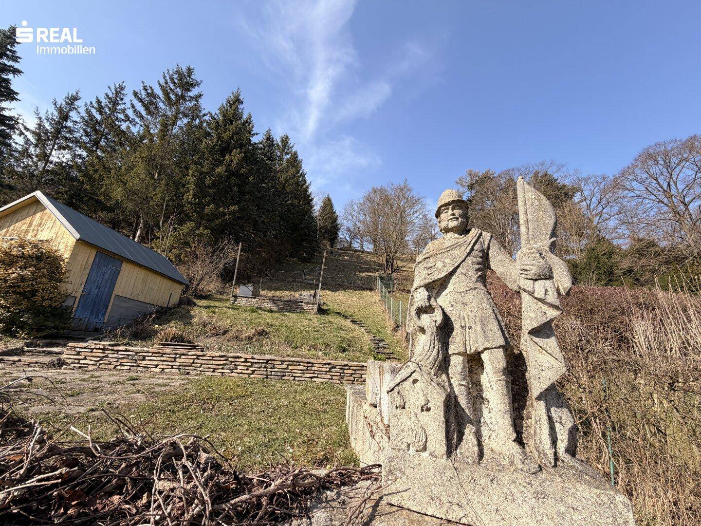 Steinstatue im weitläufigen Gartenbereich mit Holzschuppen und Treppen am Hang.