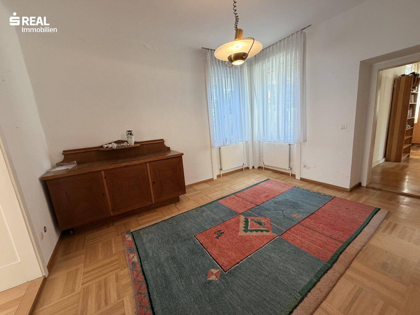 Geräumiges Zimmer mit Fischgrätparkett, großem Fenster und einem eleganten Sideboard.