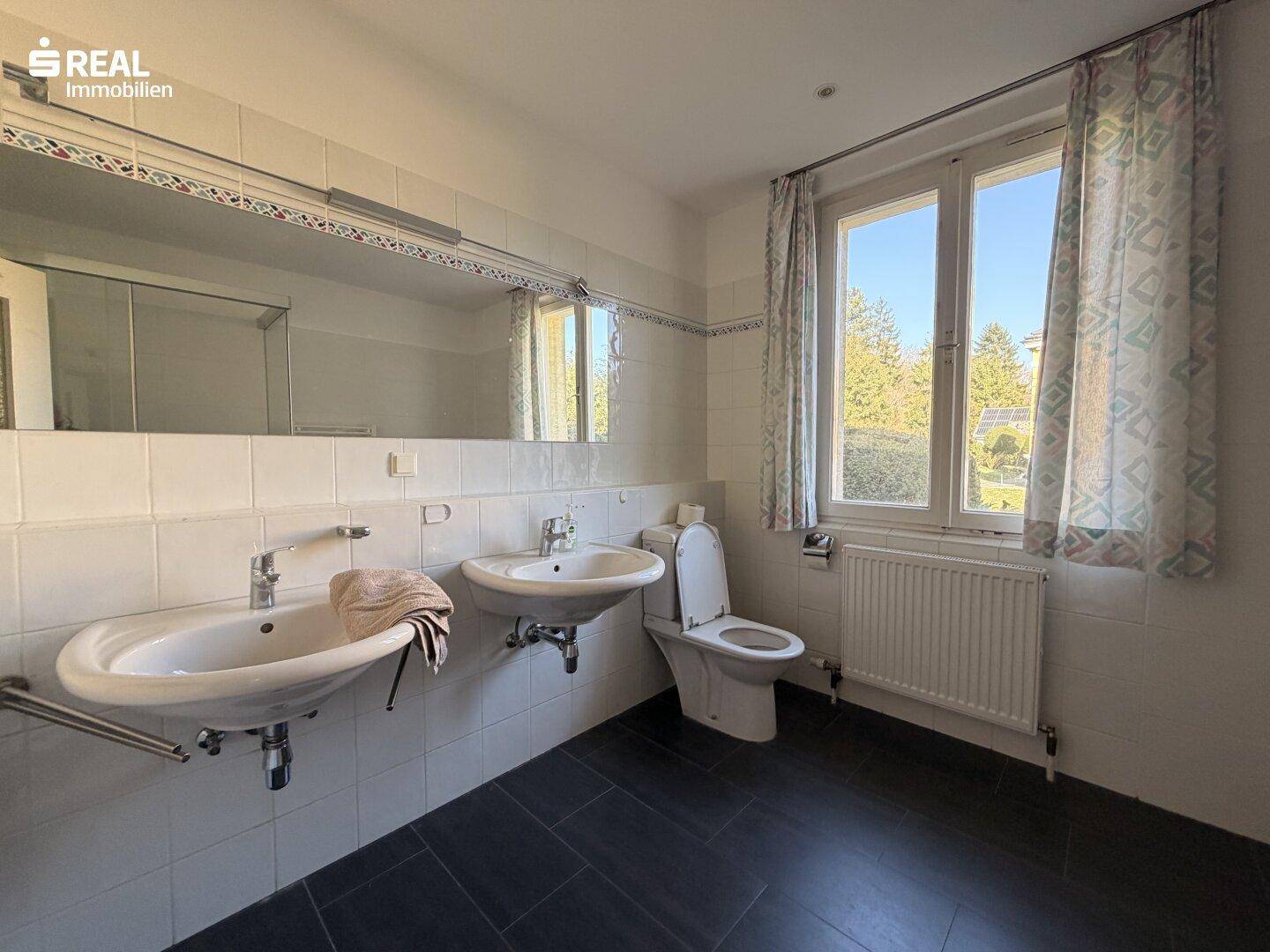 Geräumiges Badezimmer mit Doppelwaschbecken, WC und Fenster, hell gefliest.