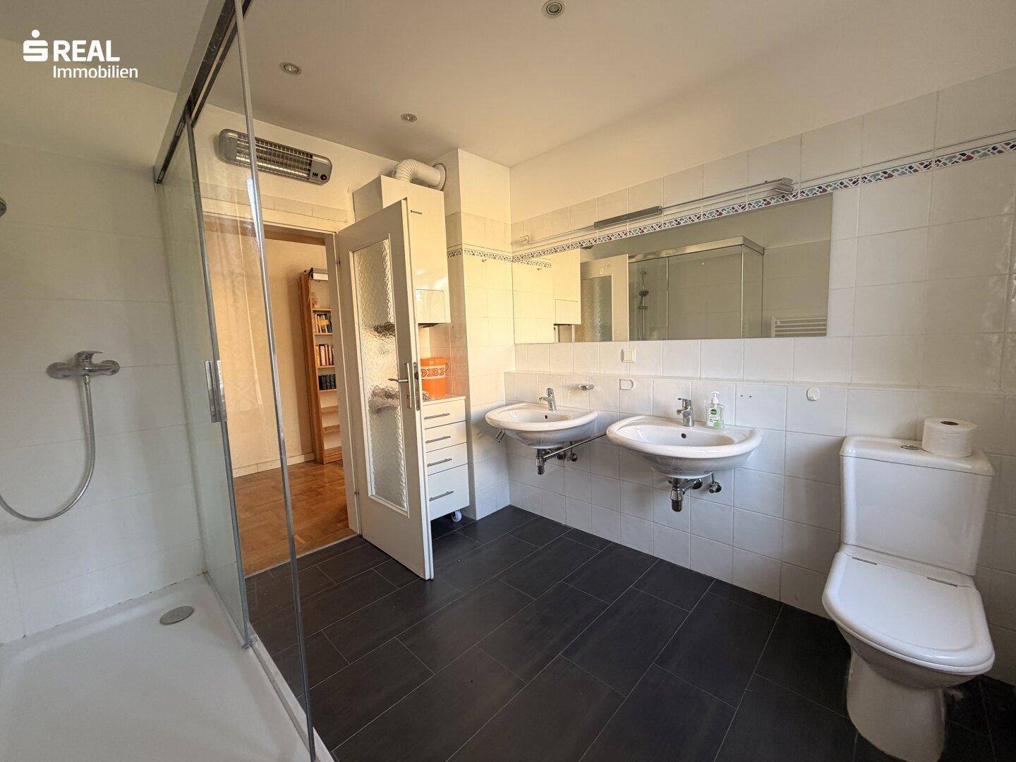 Badezimmer mit Duschkabine, Doppelwaschbecken und WC, hell gefliest.