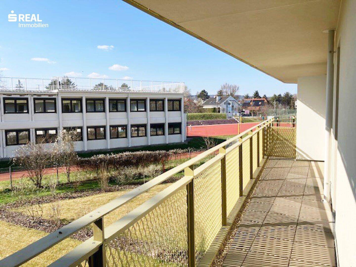 Langer Balkon mit Fliesenboden und Blick auf eine Sportanlage und Grünflächen.