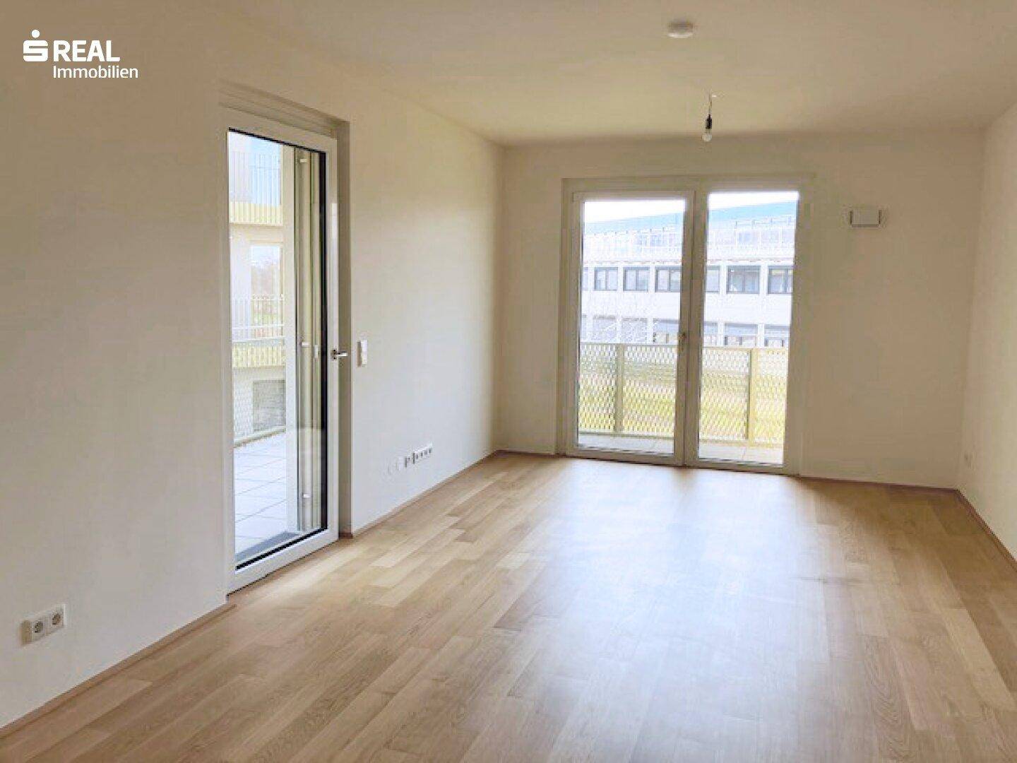 Helles, leeres Wohnzimmer mit Parkettboden und Zugang zu einem Balkon.