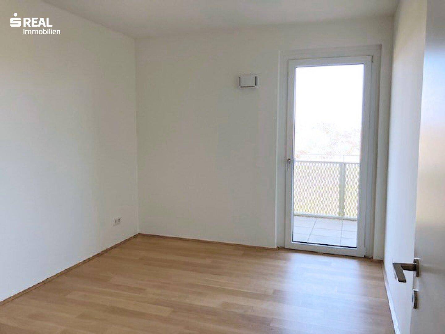 Leeres Zimmer mit Parkettboden und Zugang zu einem Balkon.