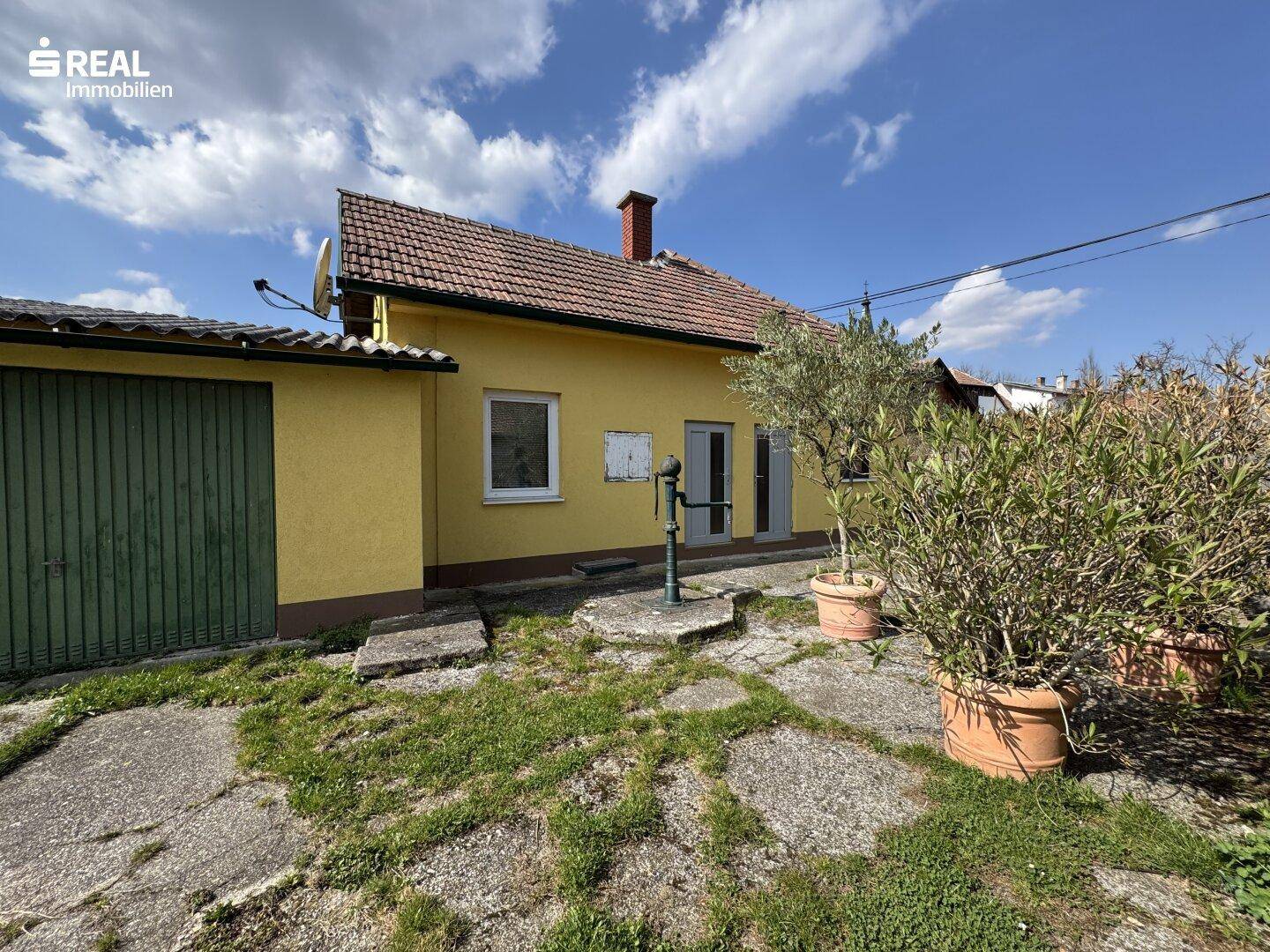 Hinterhof eines gelben Hauses mit einer Garage, Betonplatten und einer Wasserpumpe im Garten.