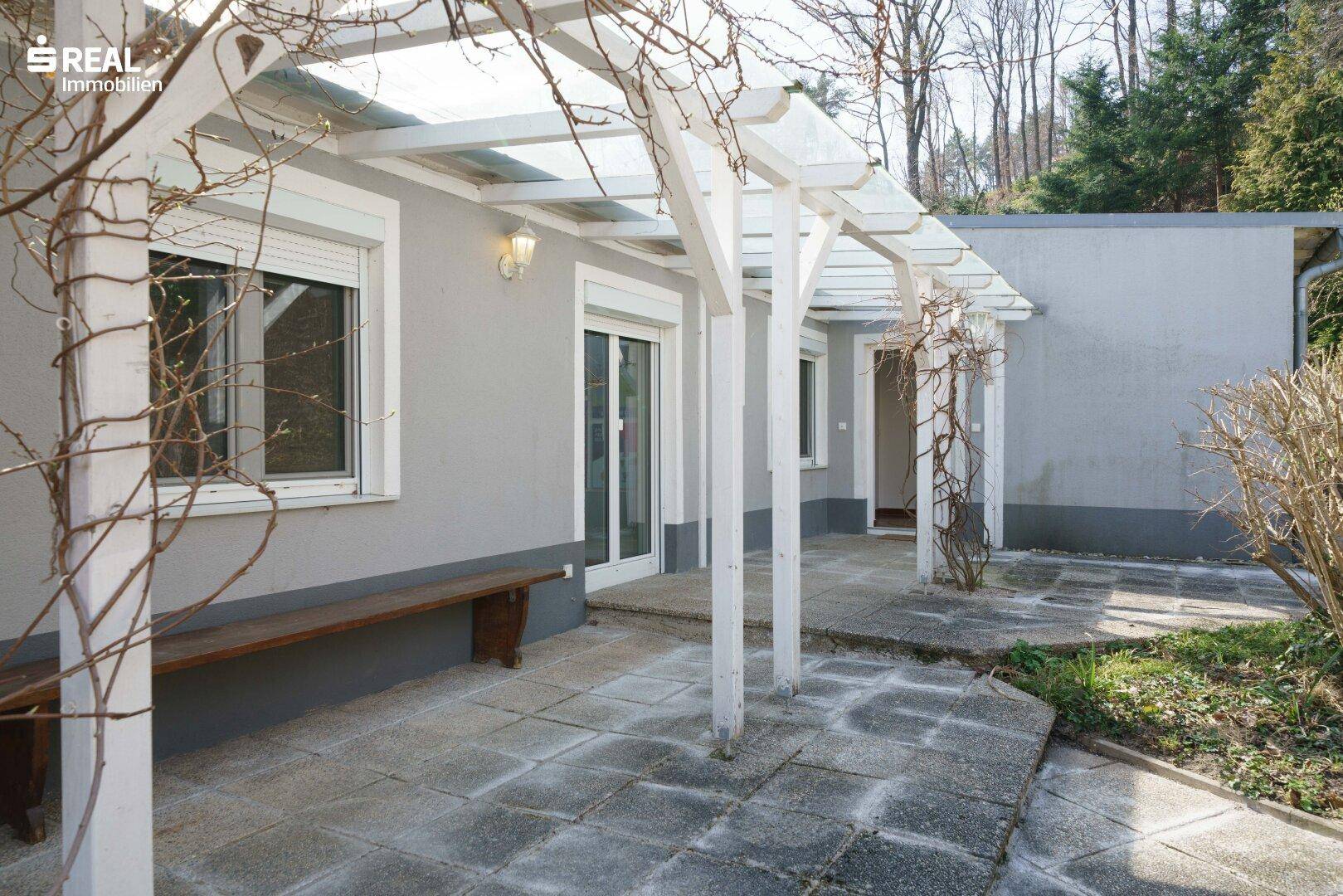 Seitliche Terrasse mit Pergola und Holzsitzbank, umgeben von Bäumen und Sträuchern.