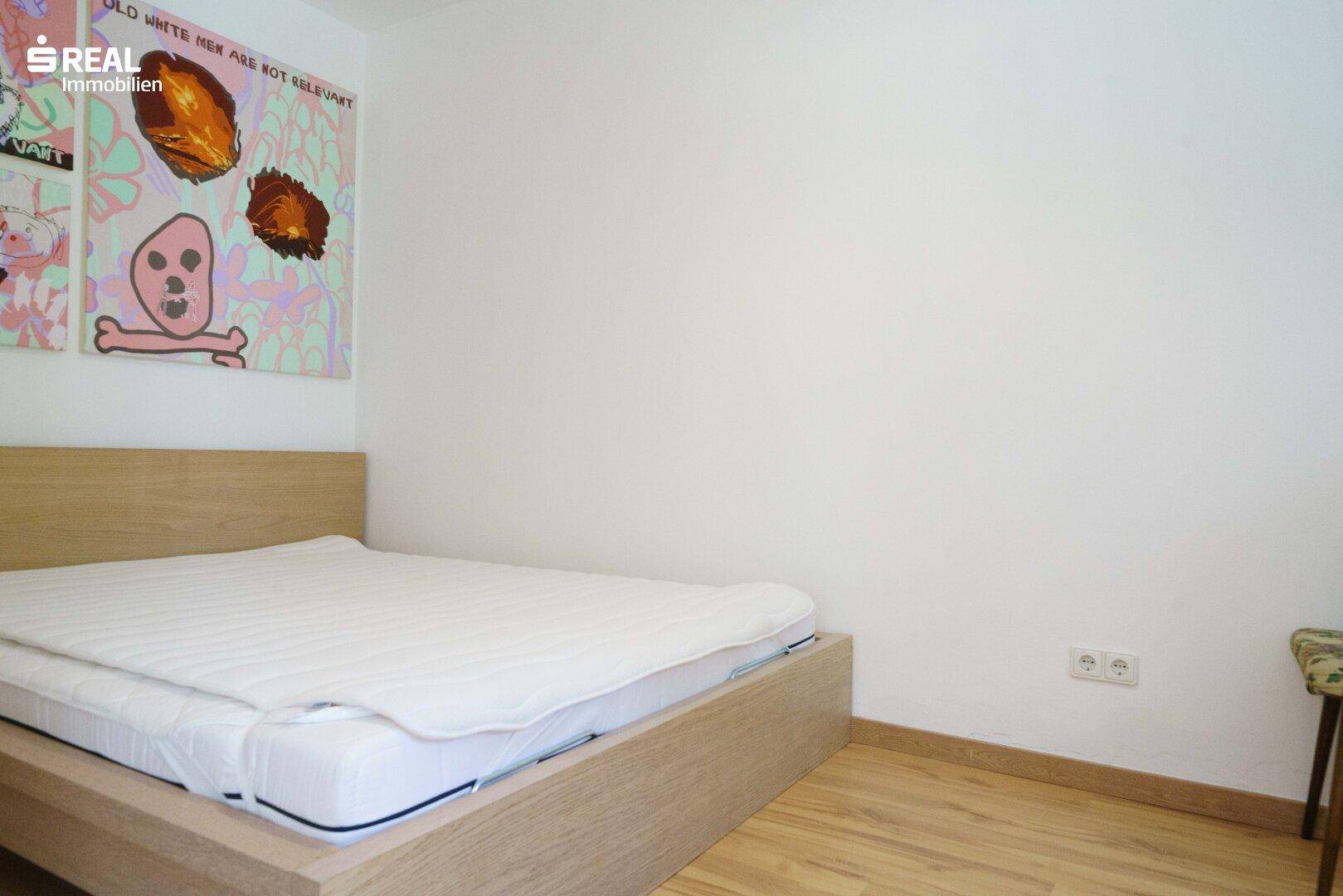 Schlafzimmer mit einfachem Bettrahmen aus Holz und hellem Laminatboden.