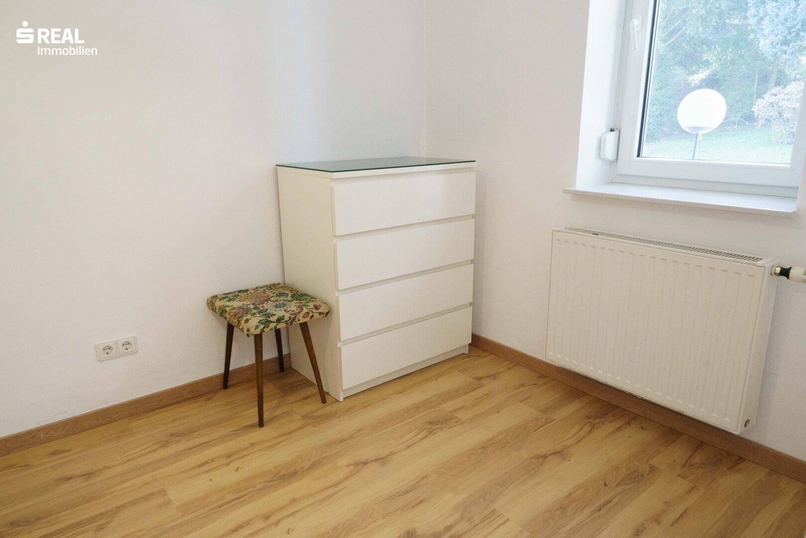 Schlafzimmer mit weißer Kommode, Heizkörper und Fenster mit Blick ins Grüne.
