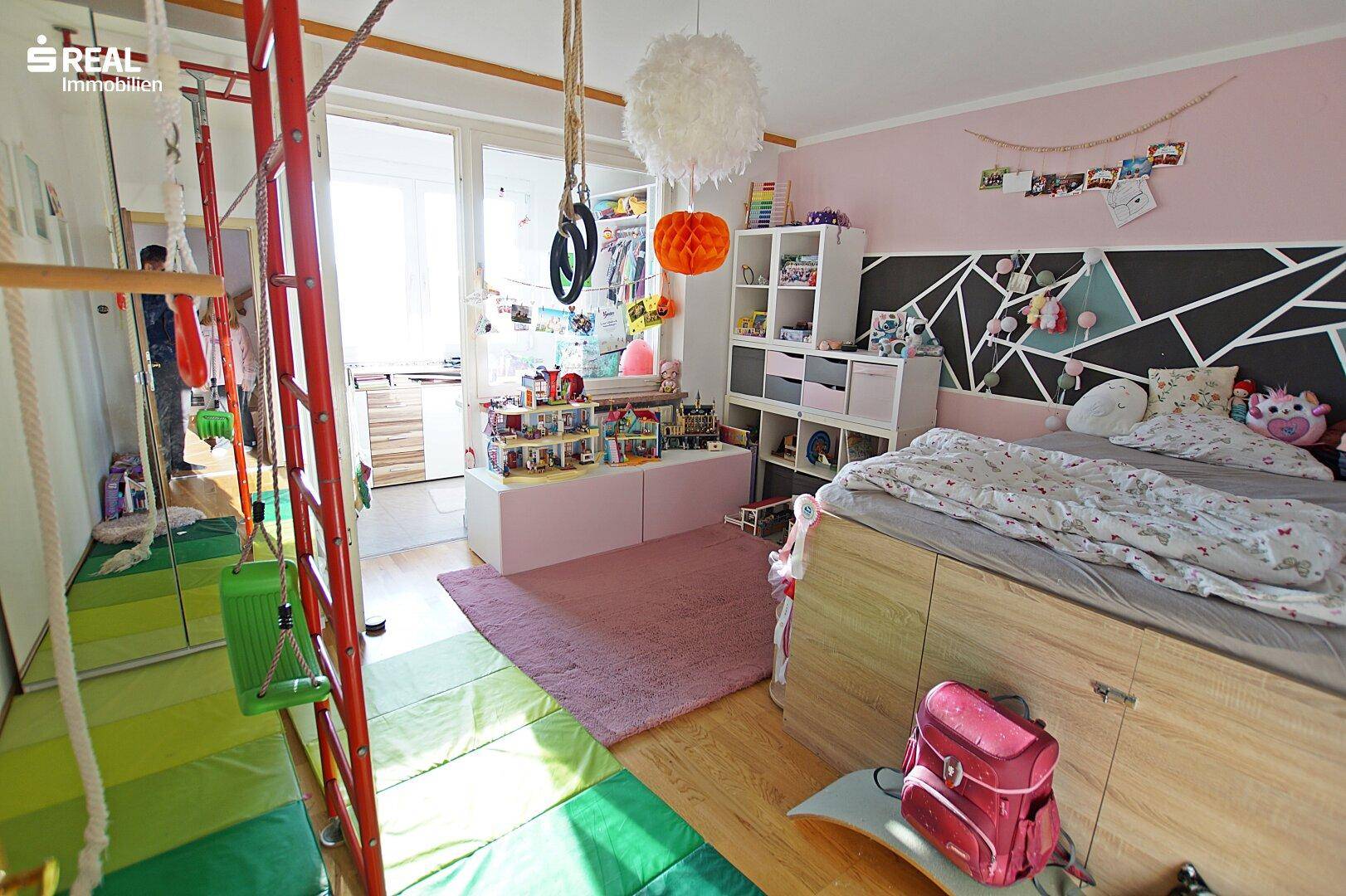 Kinderzimmer mit Spielgeräten, rosa Teppich und farbenfroher Wandgestaltung.