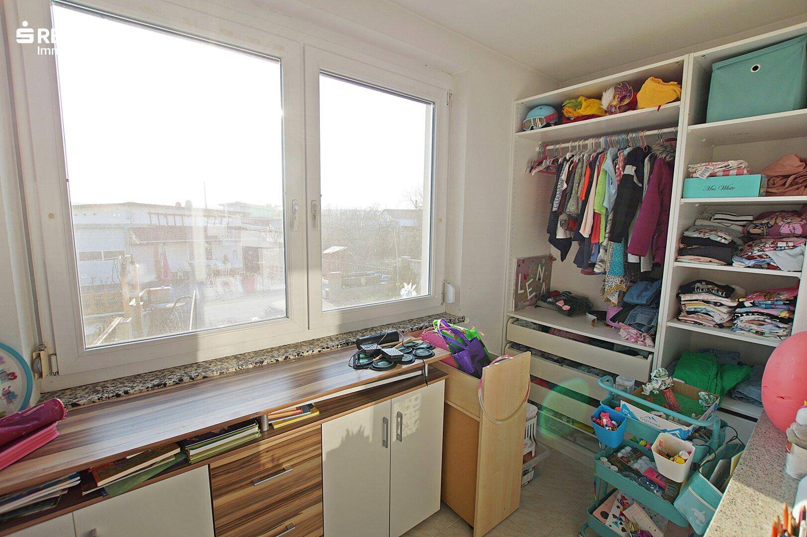 Heller Bereich im Kinderzimmer mit großem Fenster und offenem Kleiderschrank.