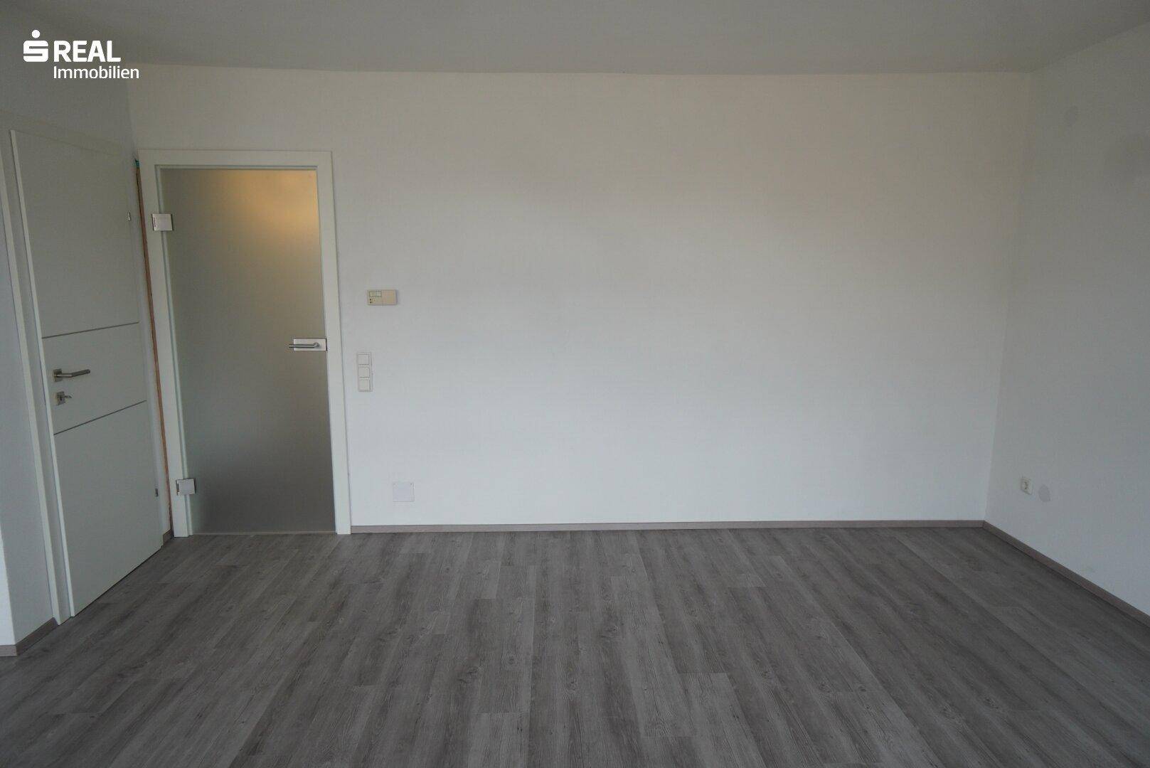 Leeres Wohnzimmer mit Laminatboden und Zugang zu weiteren Räumen.