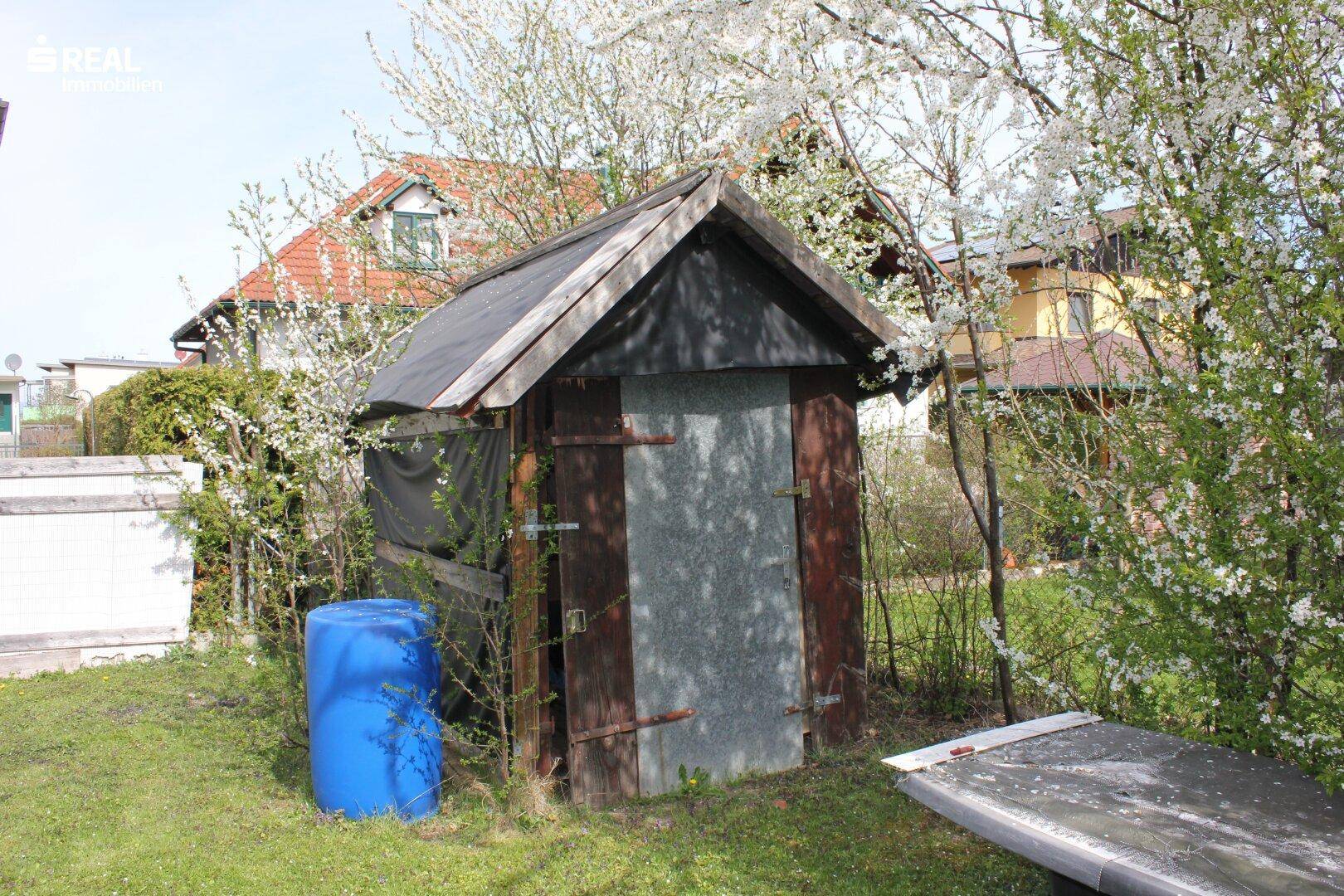 Kleiner Holzschuppen im Garten, umgeben von blühenden Sträuchern und Bäumen.