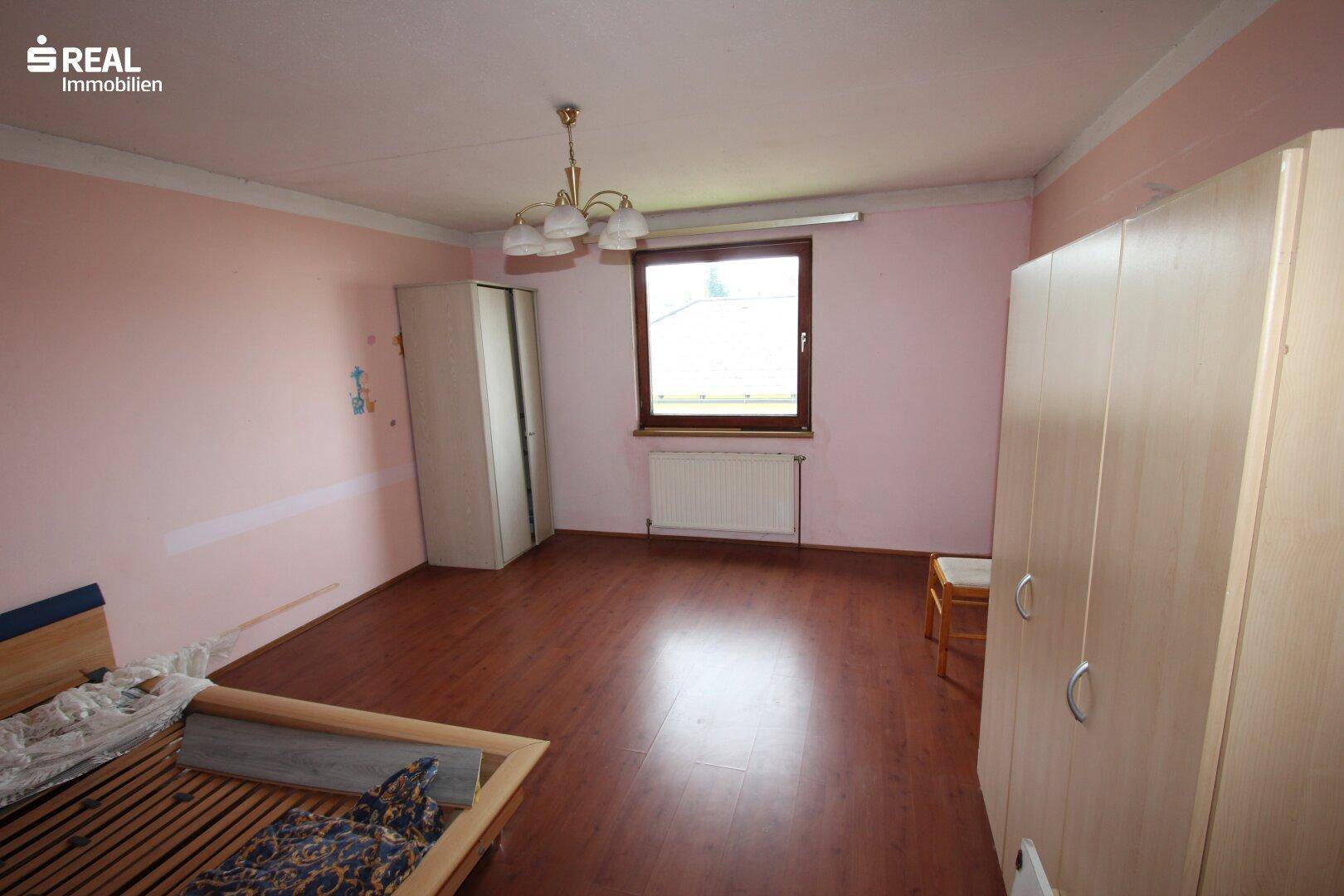 Schlafzimmer mit rosa Wänden, Holzboden und einem Fenster, das den Raum erhellt.