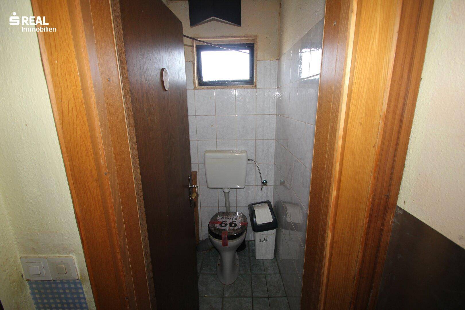Kleines WC mit weißen Fliesen, einem Fenster und einfacher Ausstattung, typisch für die 80er Jahre.