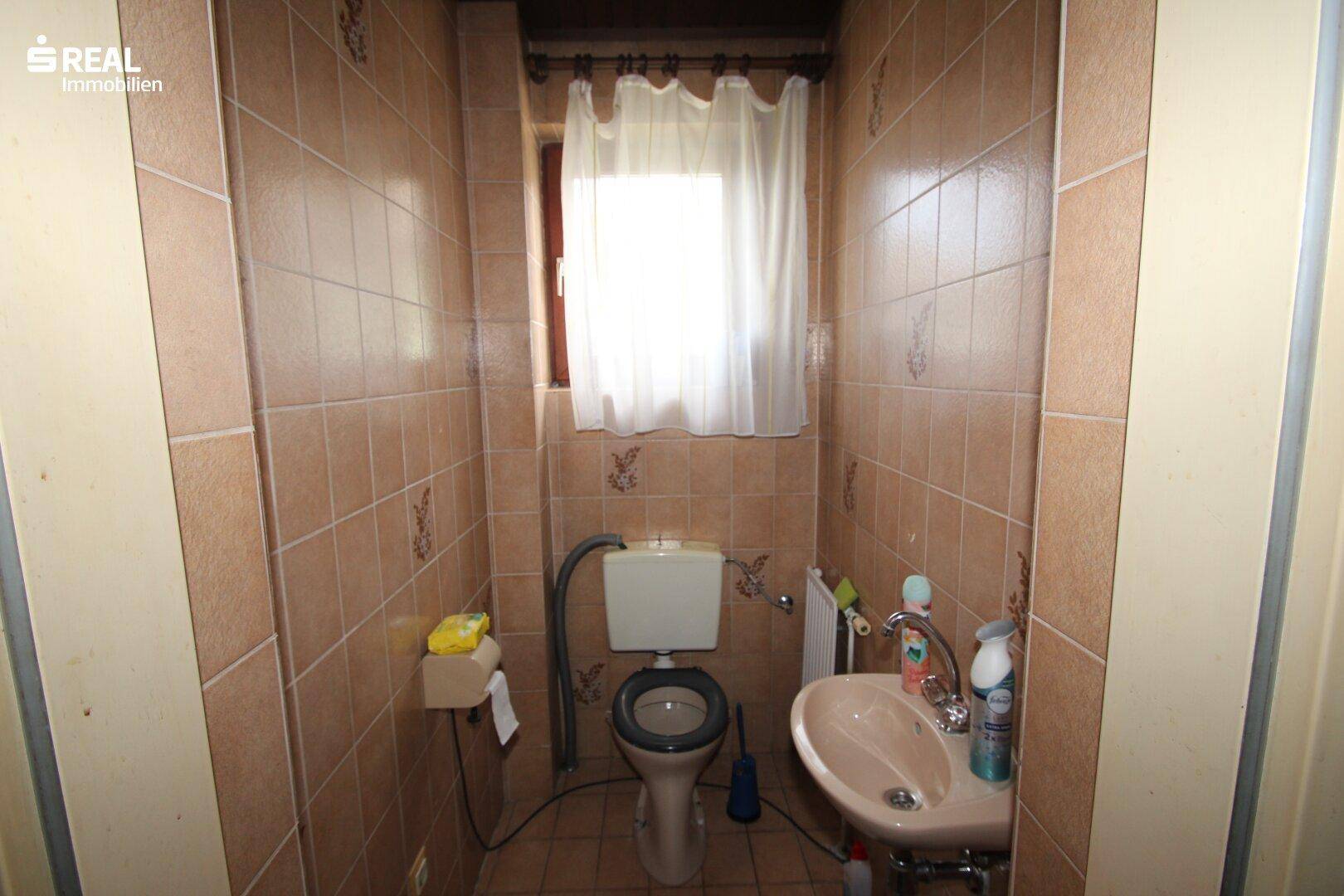 Separate Toilette mit braunen Fliesen, Fenster und einem kleinen Handwaschbecken.