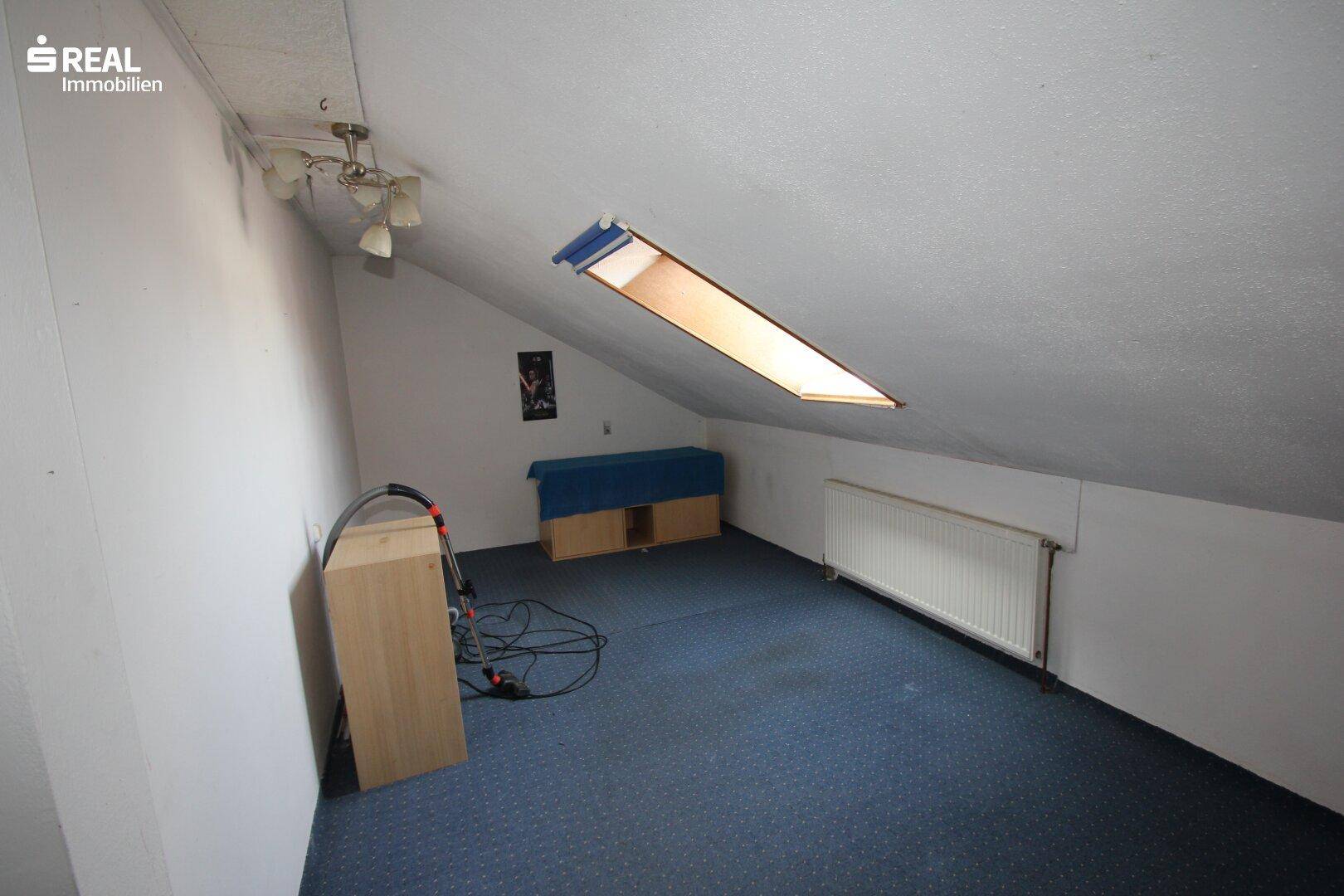 Dachgeschosszimmer mit schrägen Wänden, Dachfenster und blauem Teppichboden.