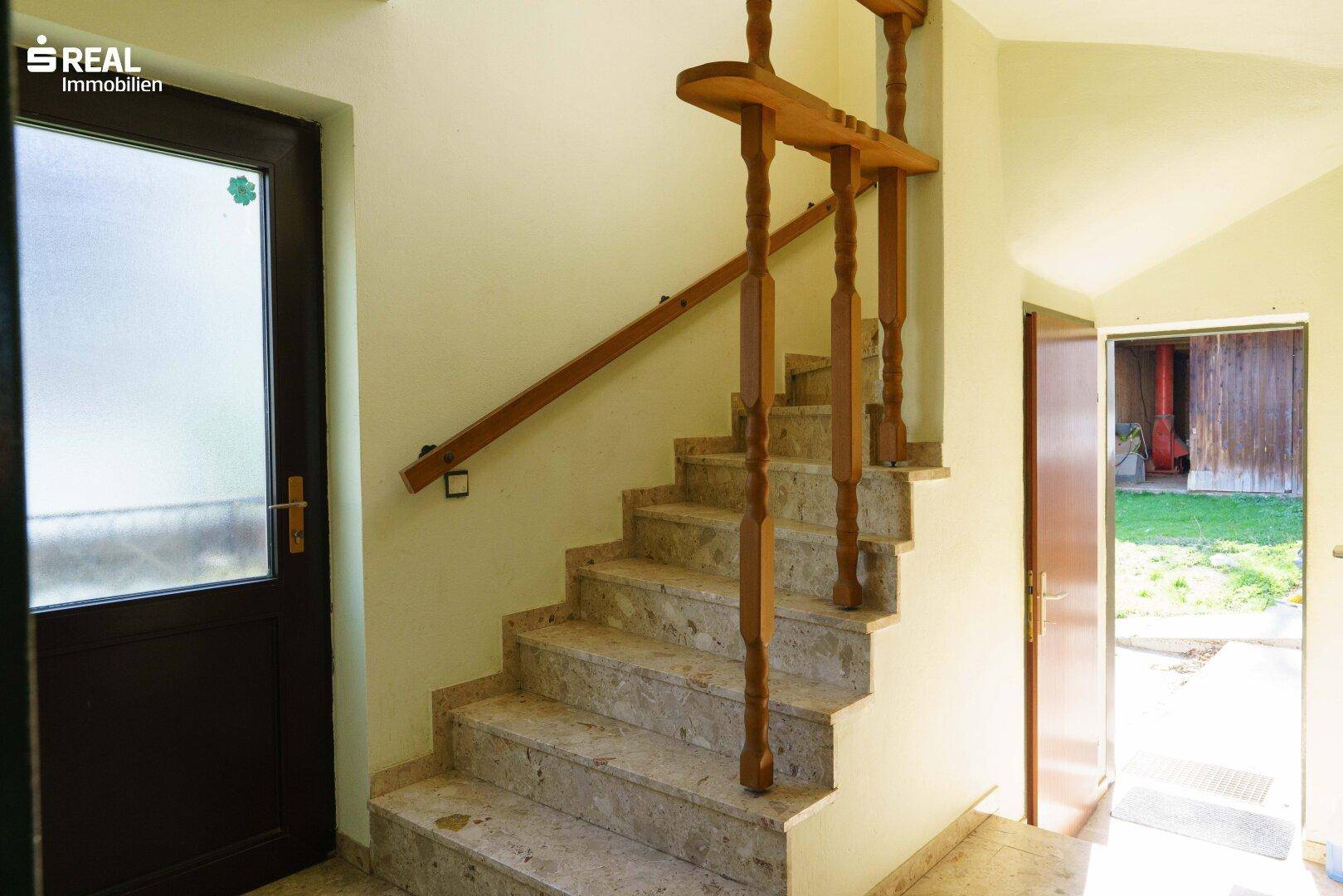Steintreppe mit Holzgeländer, die in das obere Stockwerk führt, beleuchtet von natürlichem Licht.