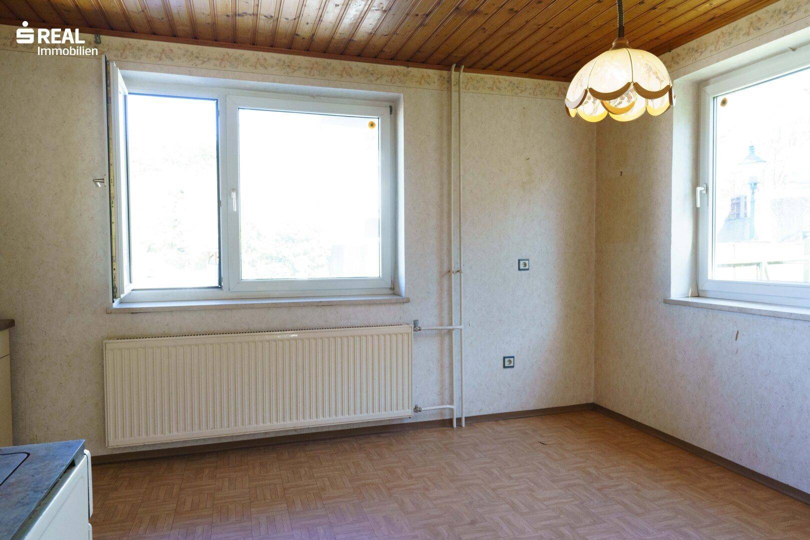 Leerer Raum mit Holzdecke, großem Fenster und Heizkörper, bereit für eine Kücheninstallation.