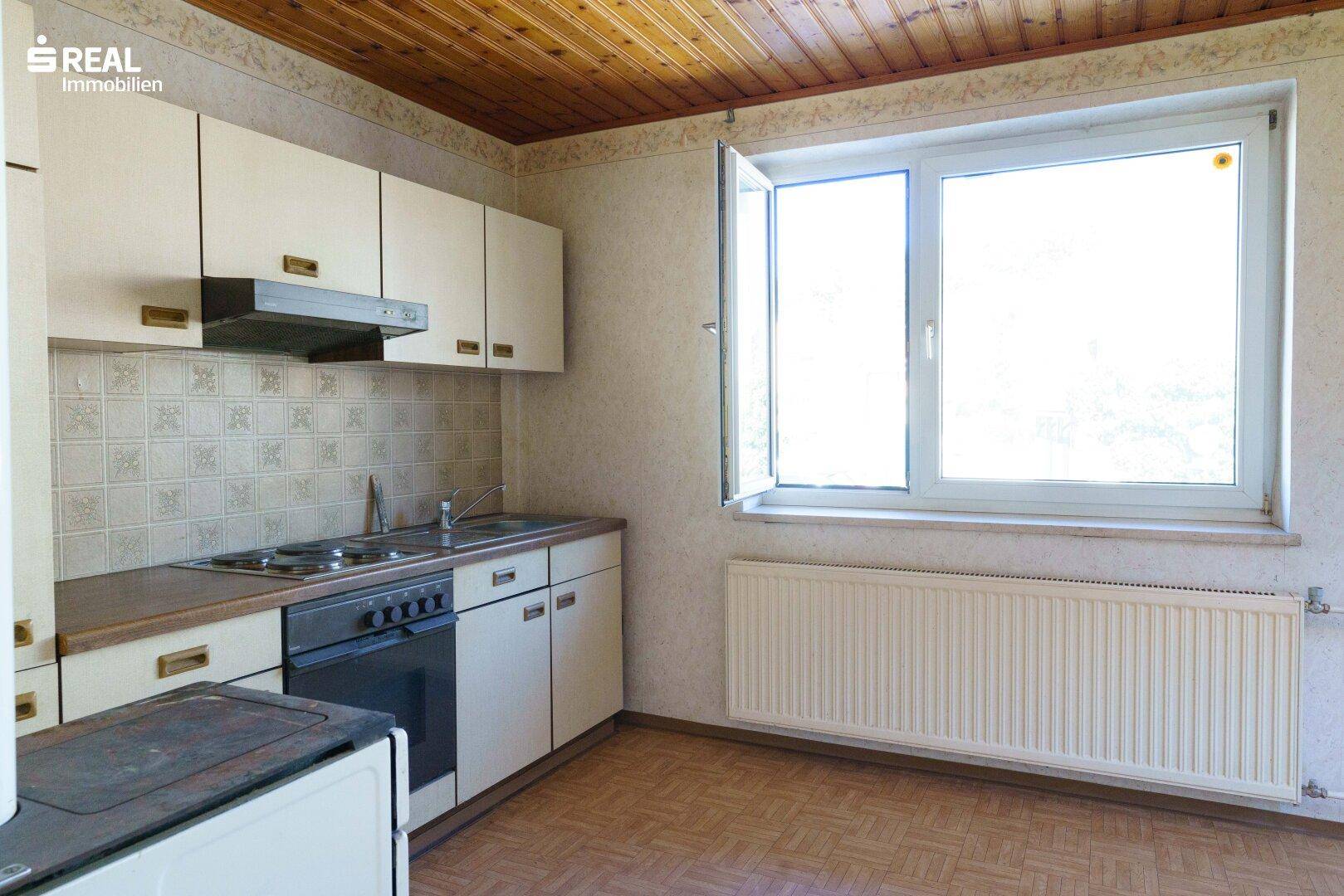 Einfache Einbauküche mit weißen Schränken, Spüle, Herd und einer Holzdecke.
