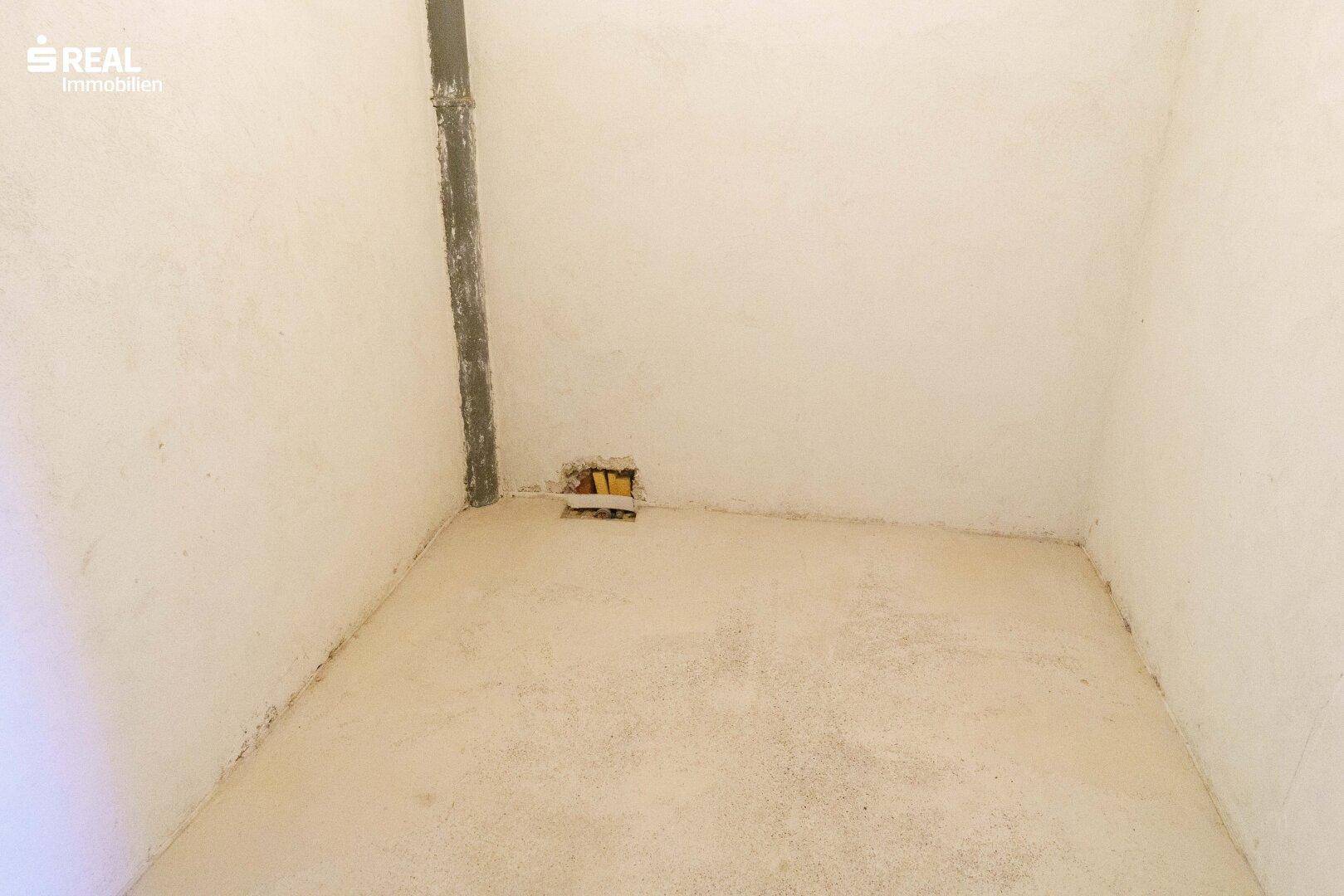 Leerer Raum mit Betonboden und einer sichtbaren Rohrleitung an der Wand.