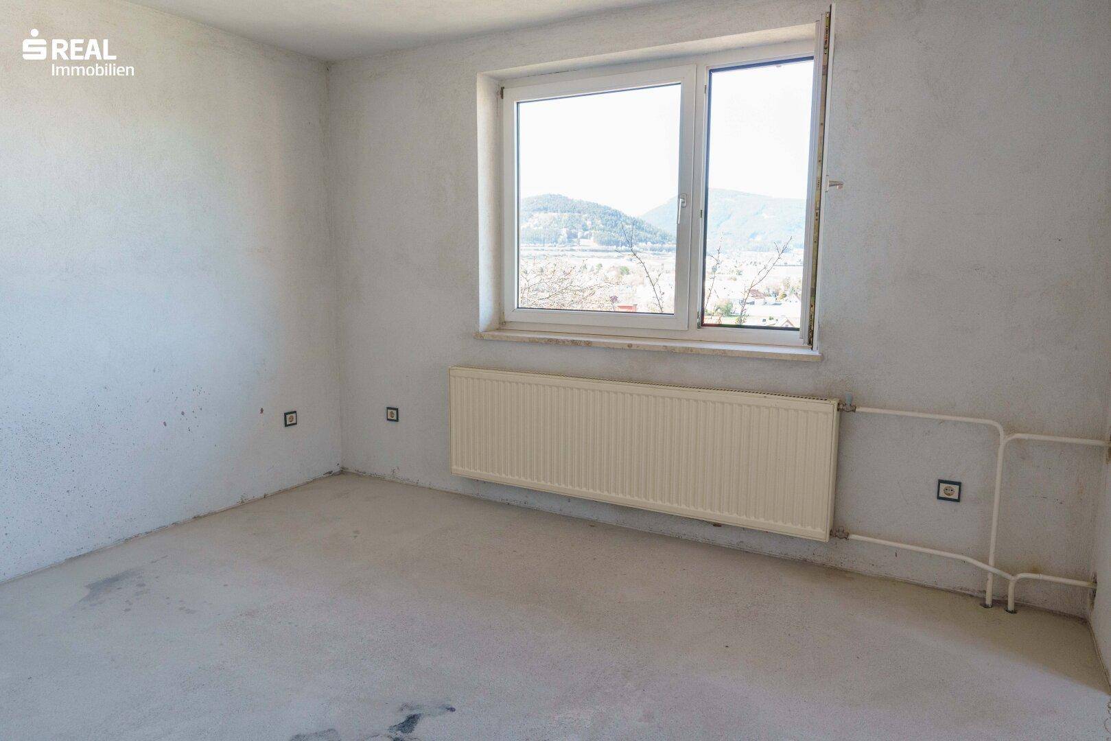 Leeres Zimmer mit Betonboden, Heizkörper und einem Fenster mit Ausblick.