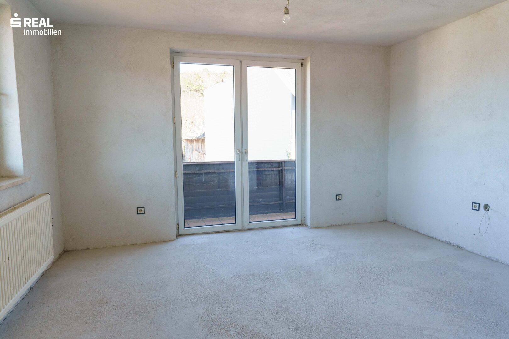 Leeres Zimmer mit Betonboden und Doppeltüren, die zu einem Balkon führen.