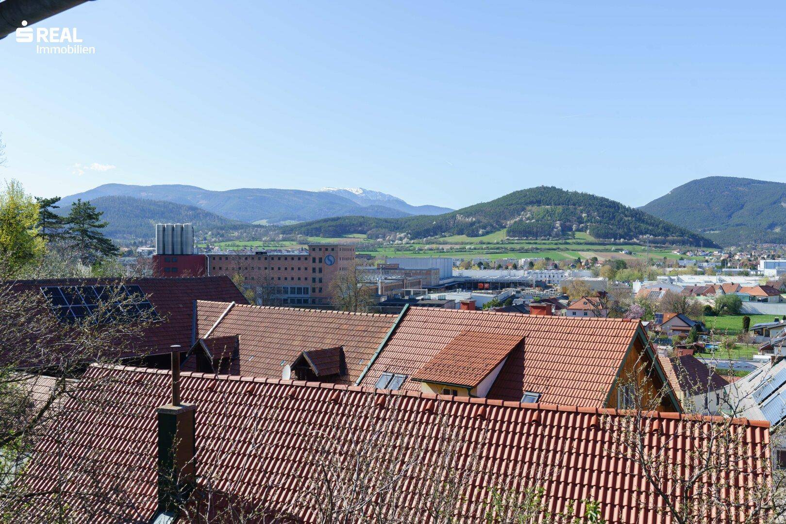 Panoramablick über die Dächer einer Stadt mit Bergen im Hintergrund unter blauem Himmel.