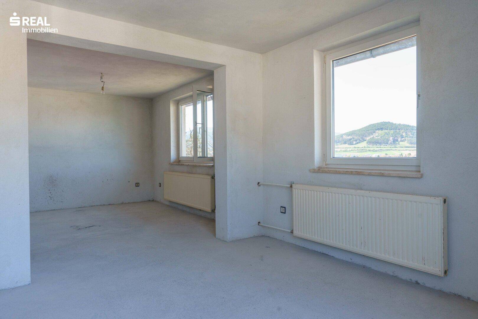 Großes, leeres Zimmer mit Betonboden und zwei Fenstern, die viel Licht hereinlassen.