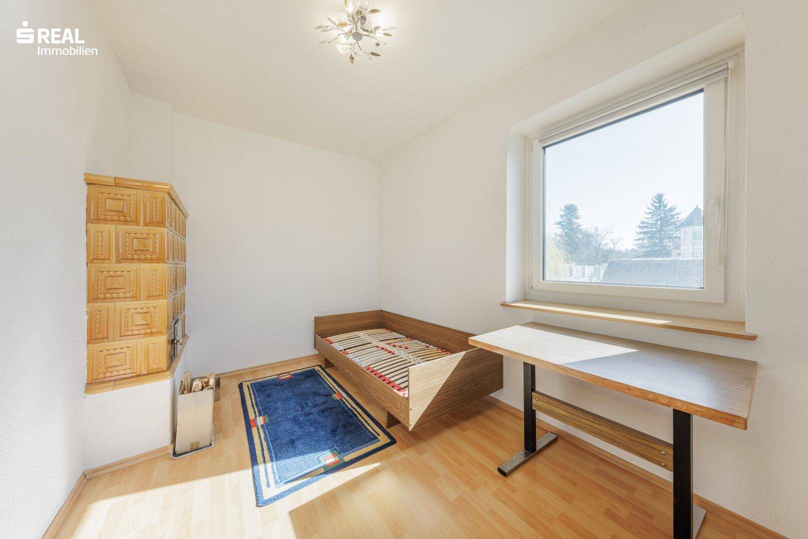 Schlafzimmer mit Kachelofen, Parkettboden und Fenster mit Blick ins Grüne.
