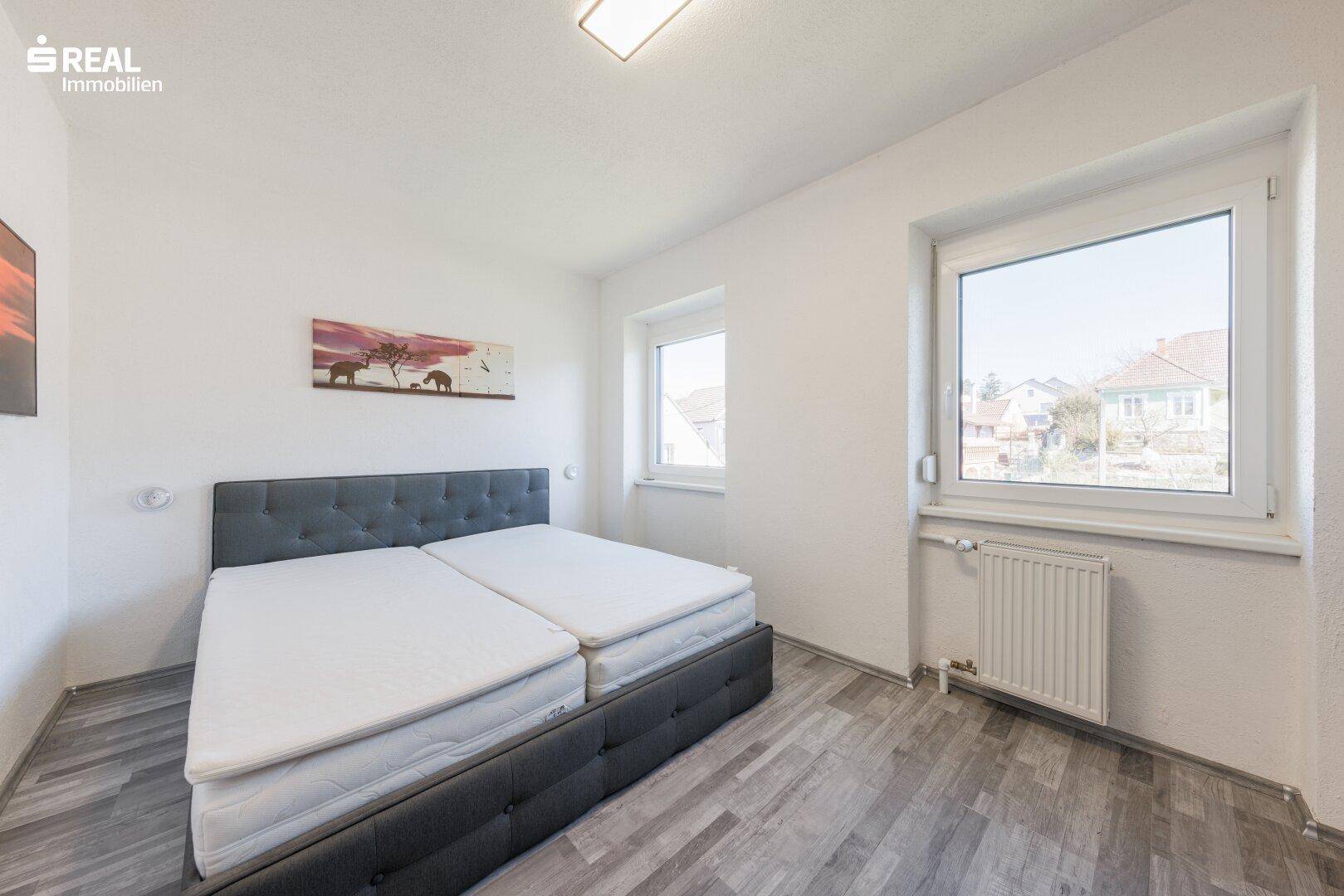 Schlafzimmer mit Doppelbett, Laminatboden und zwei hellen Fenstern.