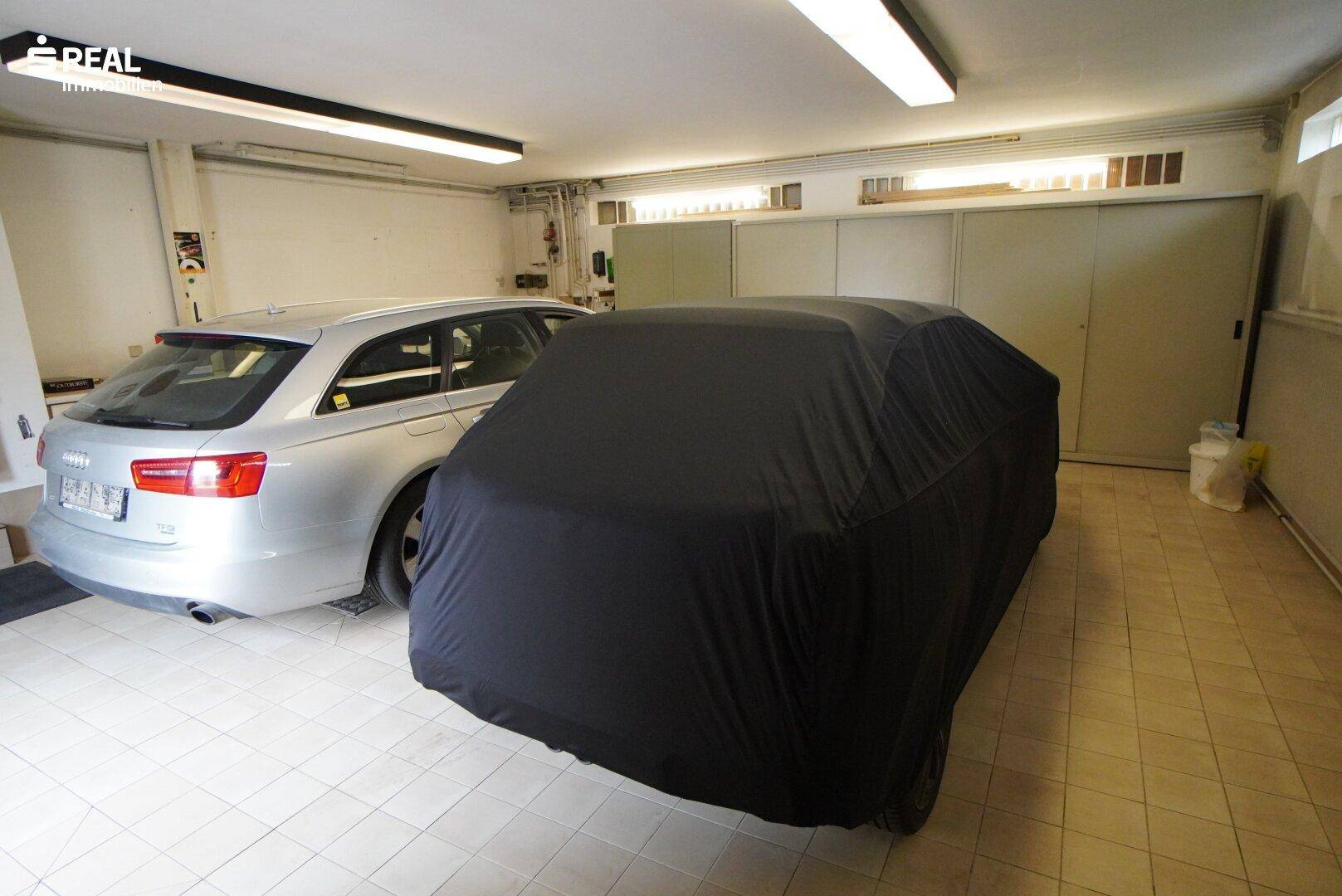 Geräumige Garage mit Fliesenboden und Platz für zwei Fahrzeuge, eines davon abgedeckt.