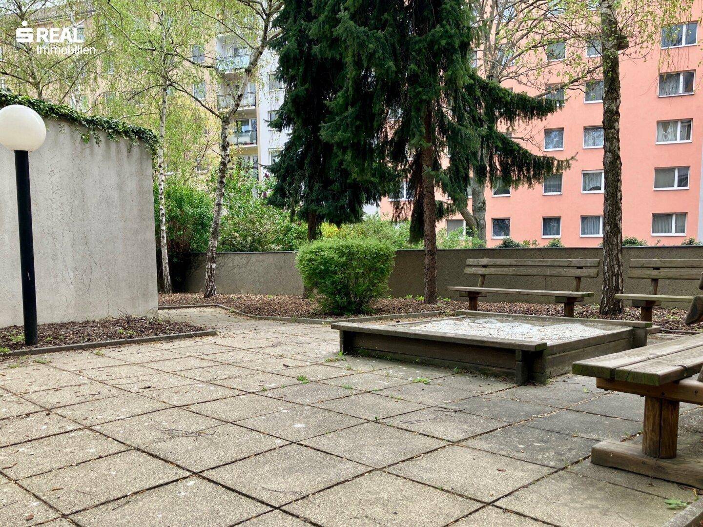Gemeinschaftsgarten mit Sandkasten, Sitzbänken und Bäumen.