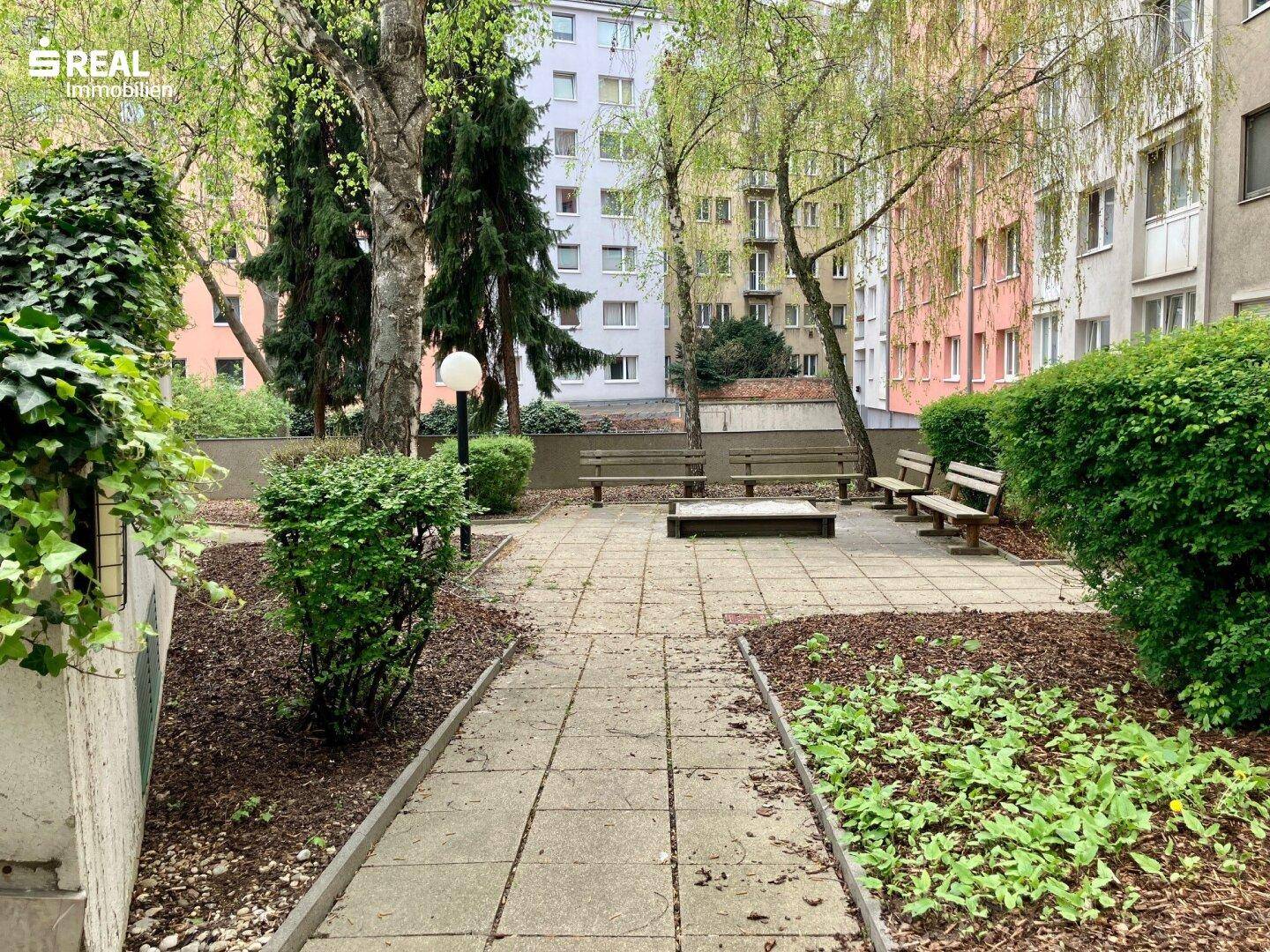 Gemeinschaftsgarten mit Sitzbänken, Bäumen und gepflasterten Wegen.