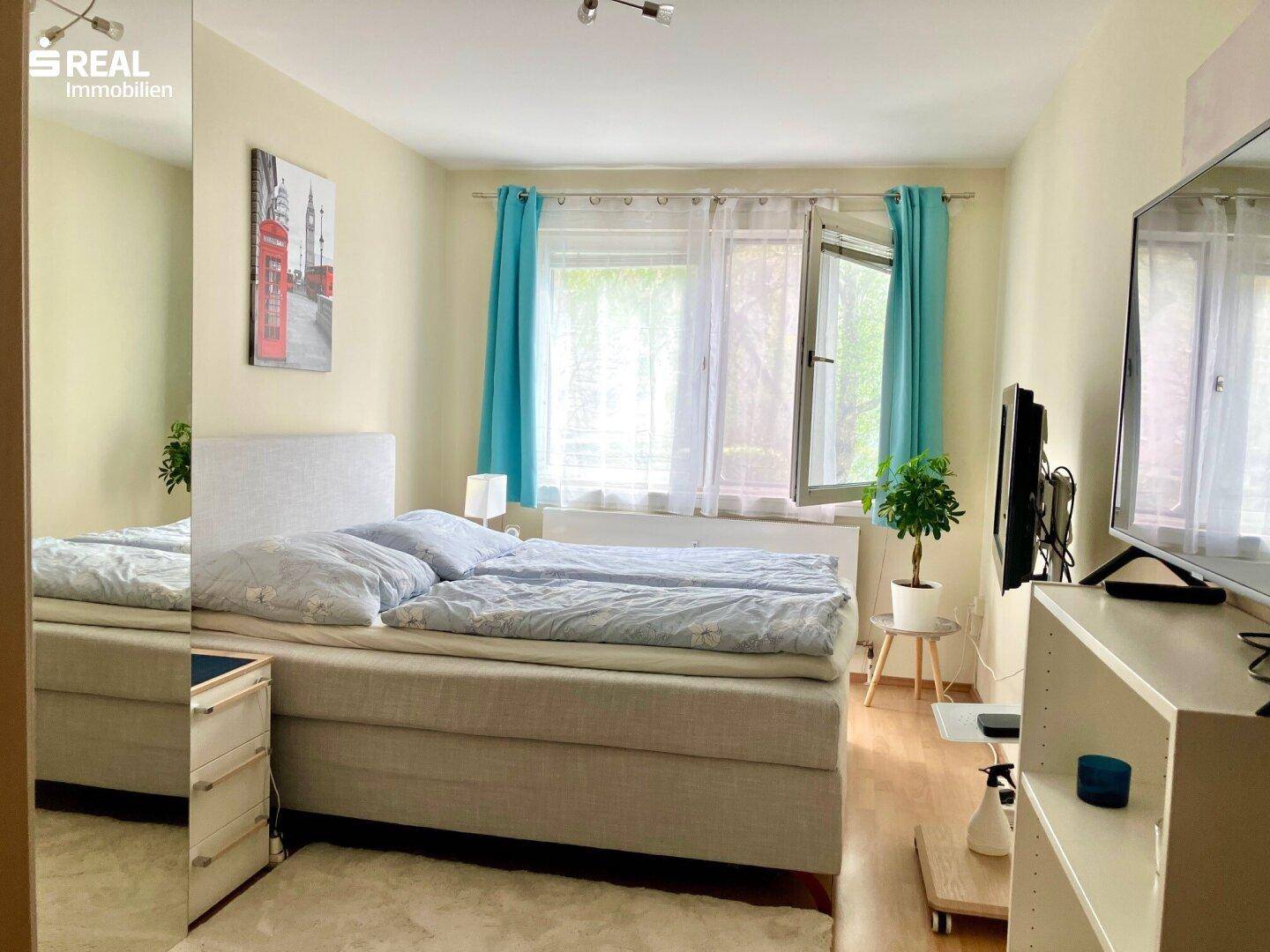 Schlafzimmer mit Doppelbett, hellem Holzboden und Fenster mit blauen Vorhängen.