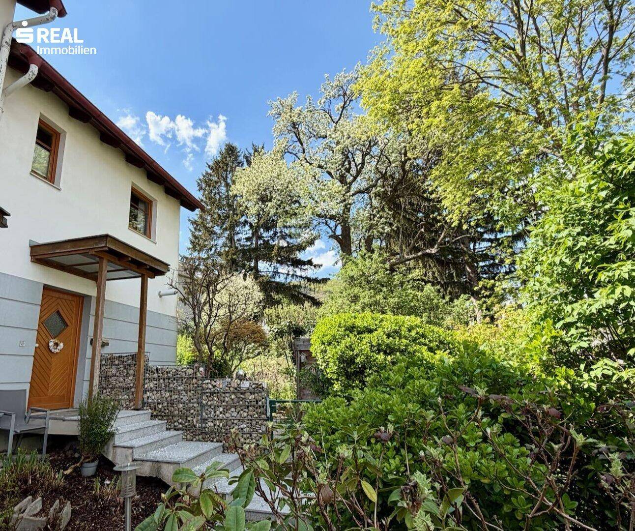 Eingangsbereich des Hauses mit Steintreppe und umgebender grüner Bepflanzung und Bäumen.