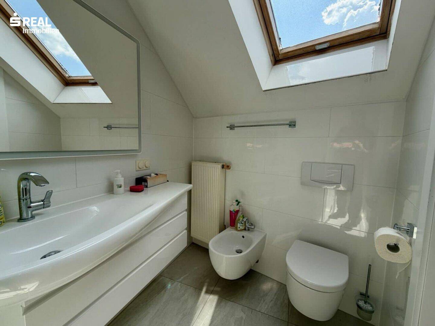 Modernes Badezimmer mit Waschtisch, Bidet, WC und Dachfenster für natürliches Licht.