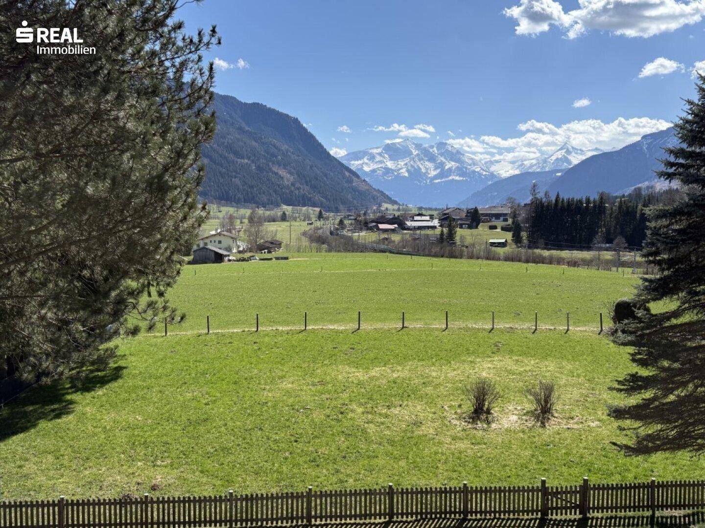 Panoramablick über grüne Felder und Wiesen mit majestätischen Bergen im Hintergrund.