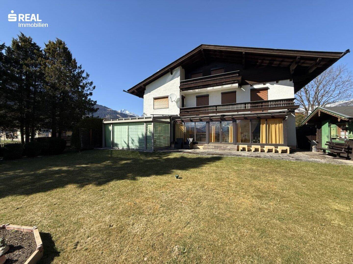 Hausfassade mit großem Garten und Wintergarten, umgeben von grüner Landschaft.