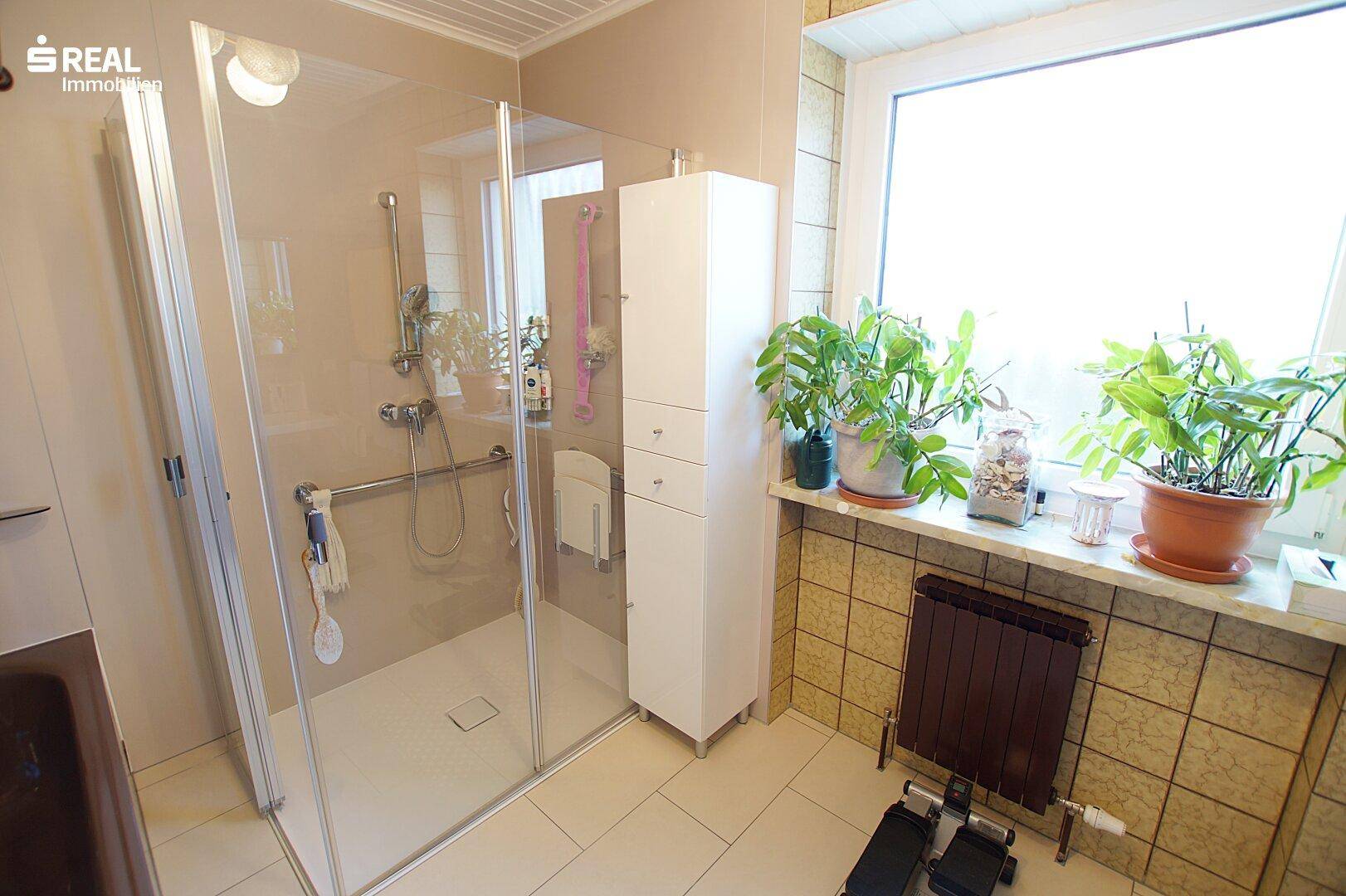 Modernes Badezimmer mit ebenerdiger Dusche, Haltegriffen und Fenster, hell gefliest.