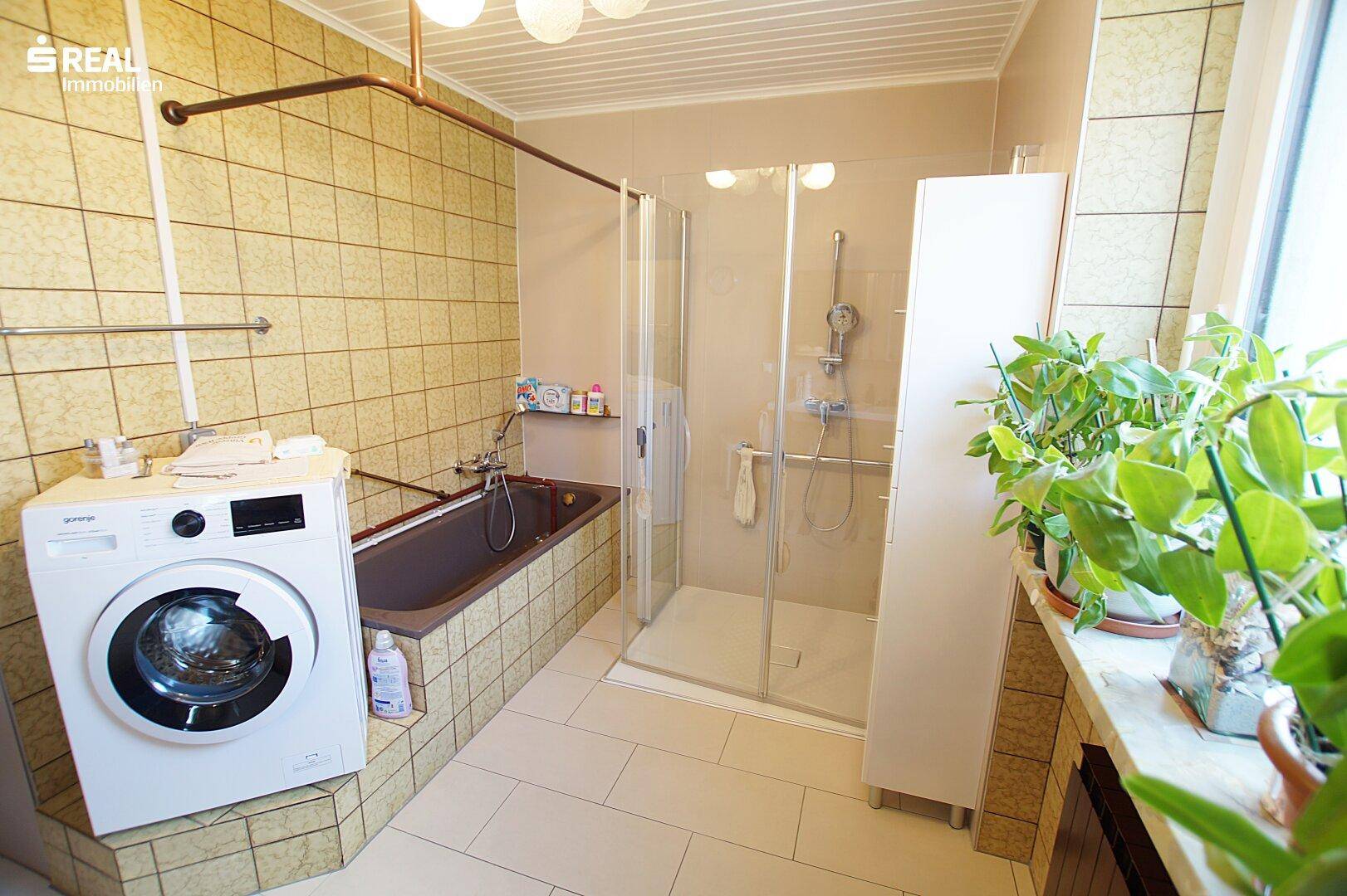 Badezimmer mit Badewanne, Dusche und Waschmaschine, hell gefliest und mit Fenster.