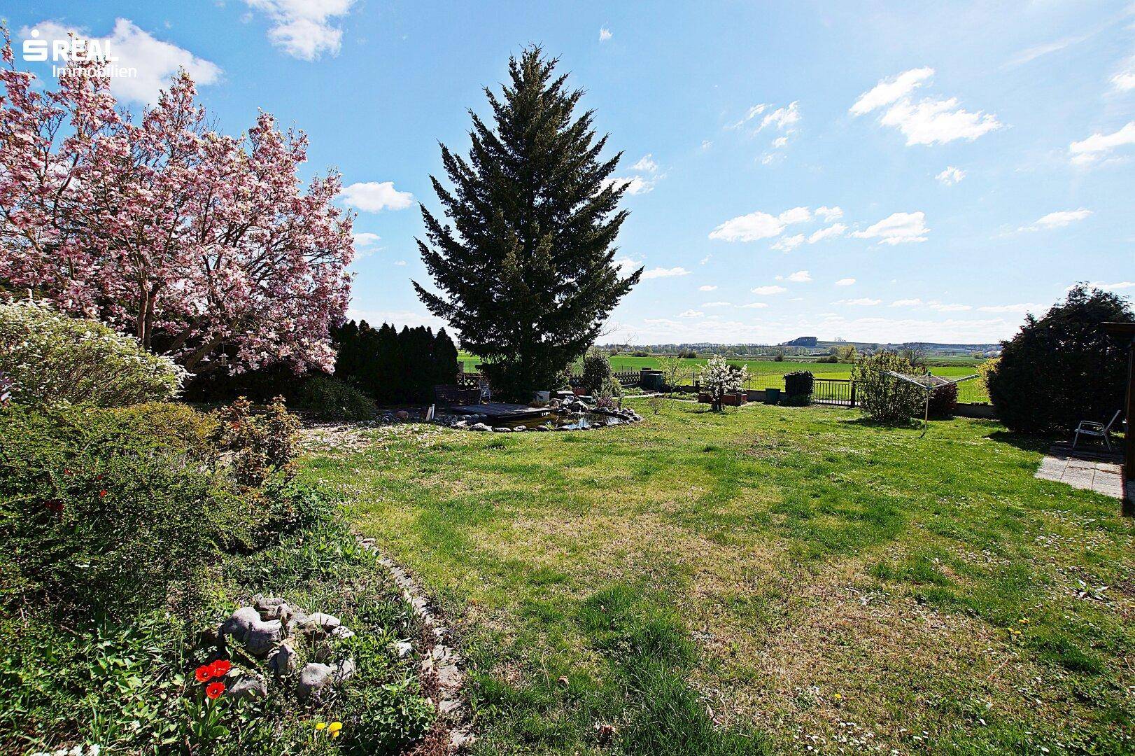Weitläufiger Garten mit großem Nadelbaum, Teich und Blick auf die umliegende grüne Landschaft.