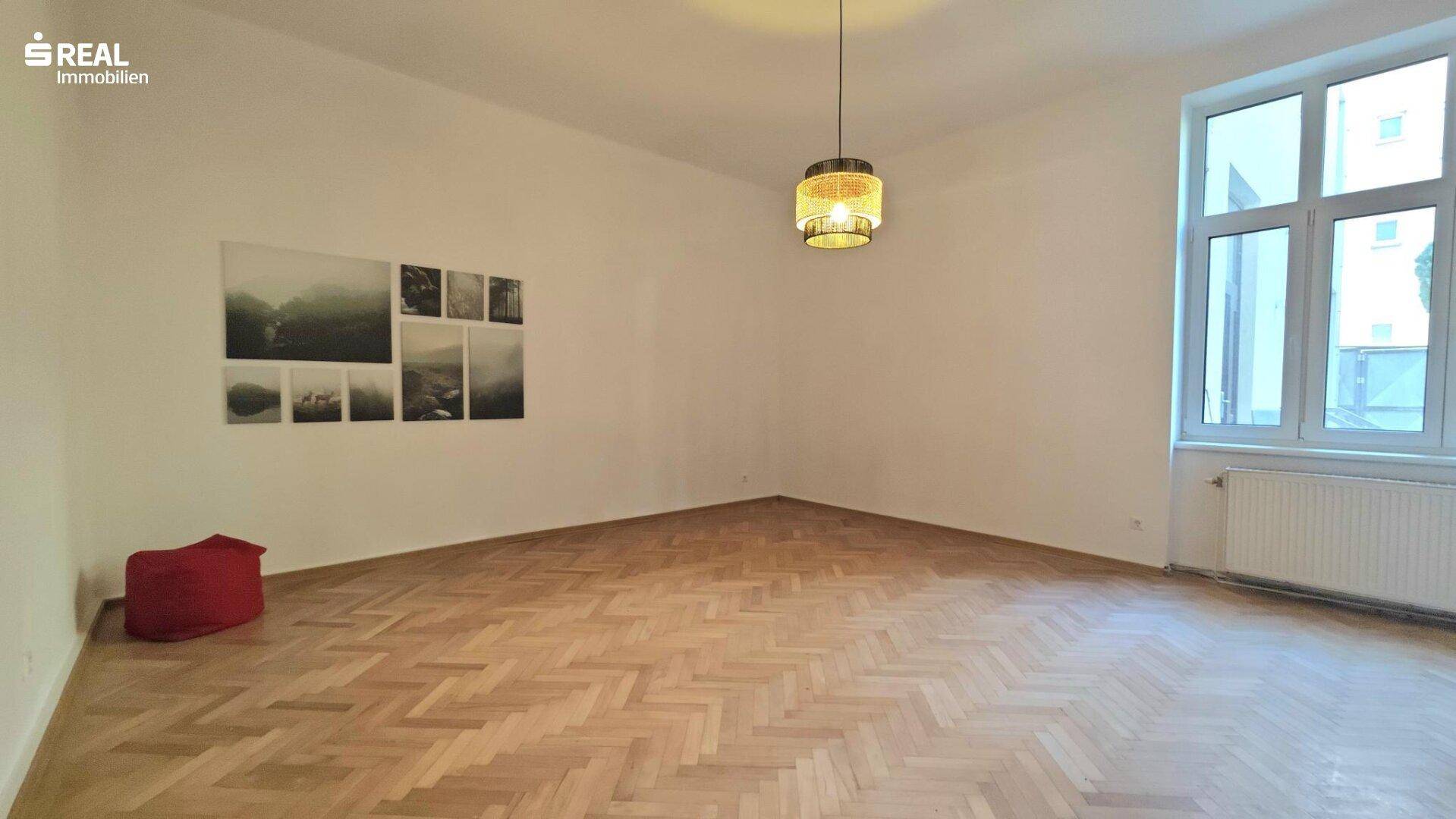 Geräumiges Wohnzimmer mit klassischem Fischgrätparkett und viel Tageslicht durch das große Fenster.