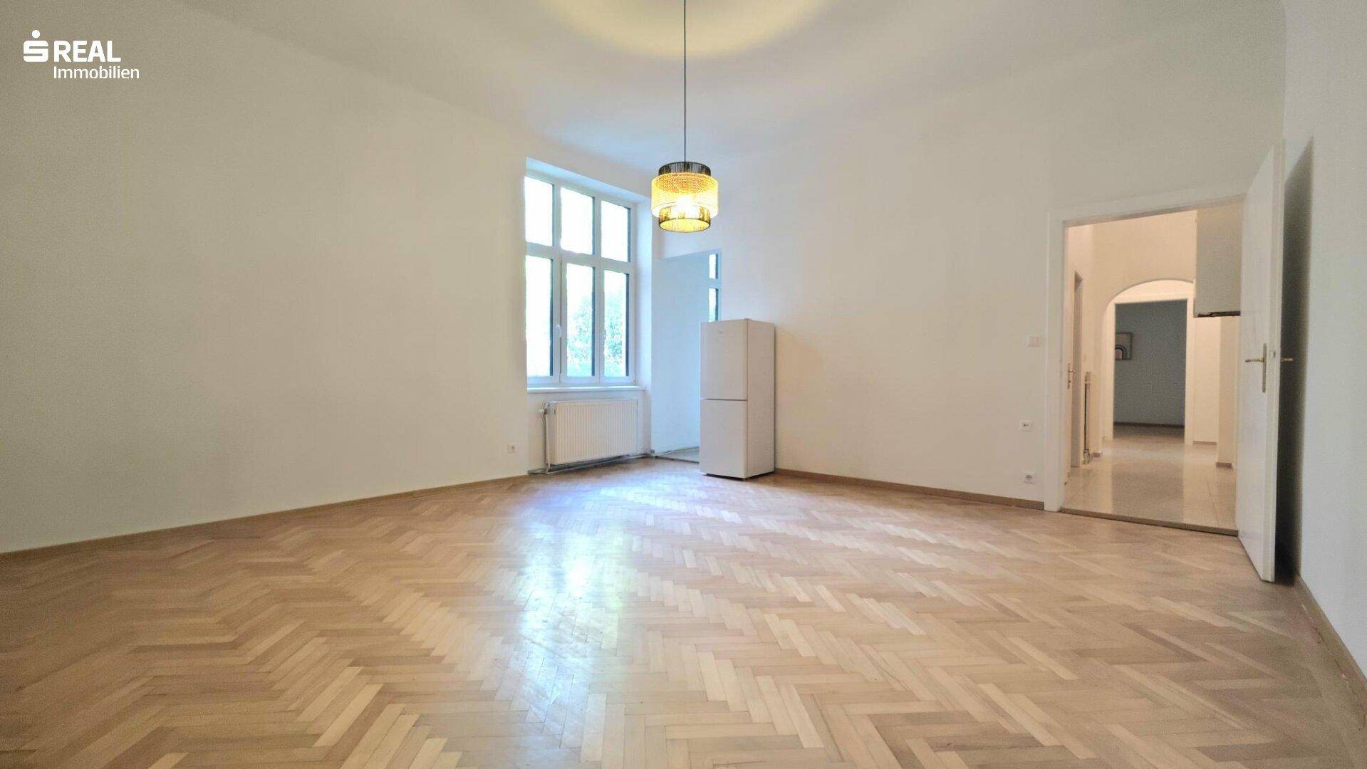 Helles Wohnzimmer mit Fischgrätparkettboden, hohen Decken und großem Fenster.