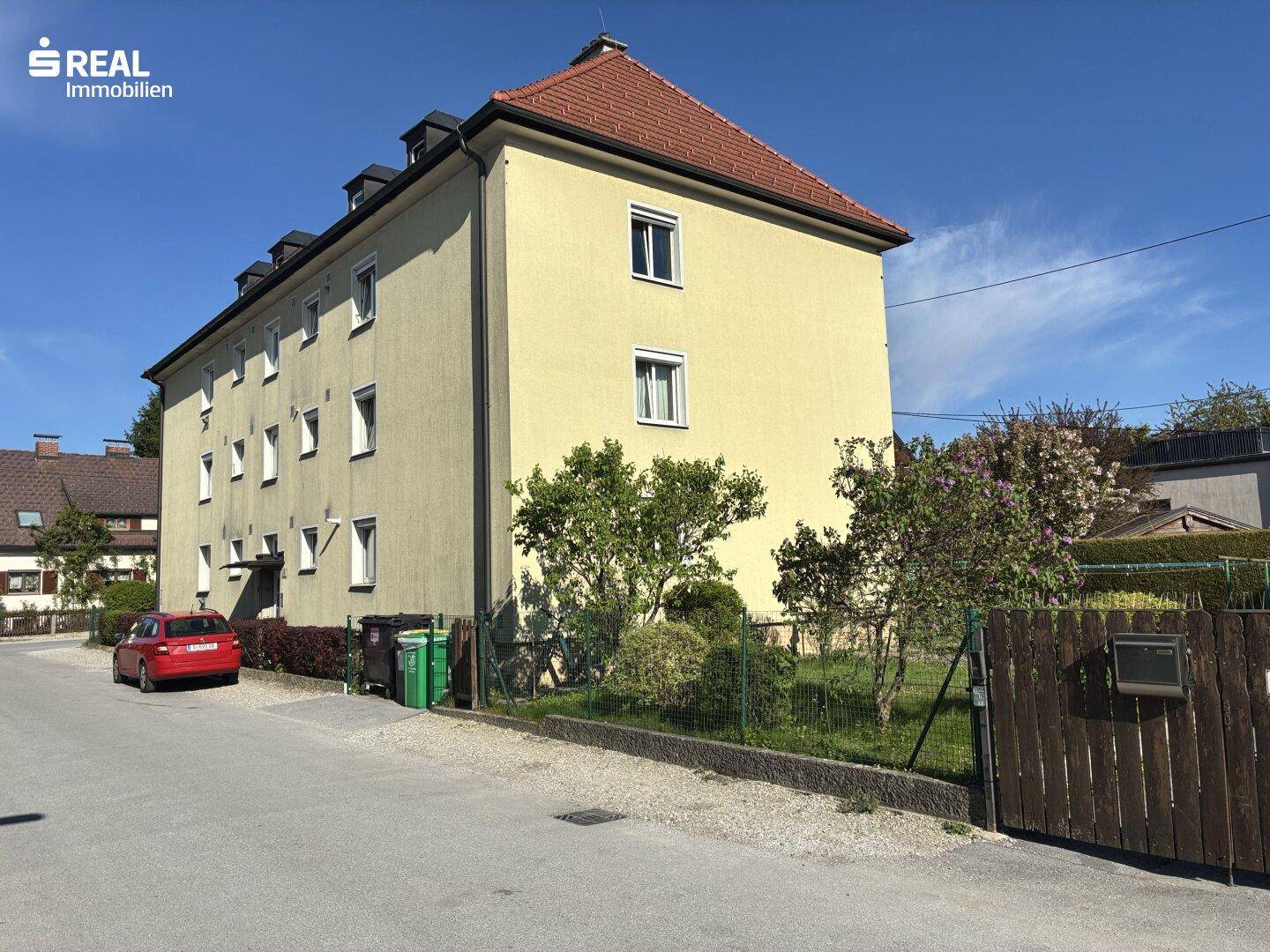 Gelbe Fassade eines Mehrfamilienhauses mit rotem Ziegeldach und kleinen Bäumen im Vorgartenbereich.